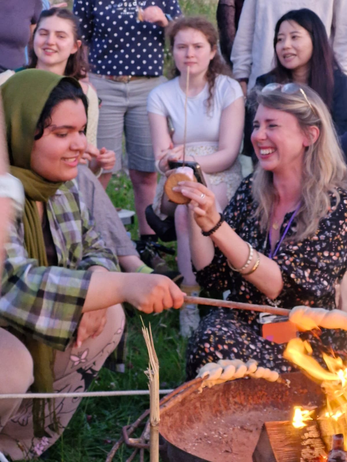 A group of women gathered outdoors around a fire pit at dusk, roasting marshmallows and enjoying each other's company.