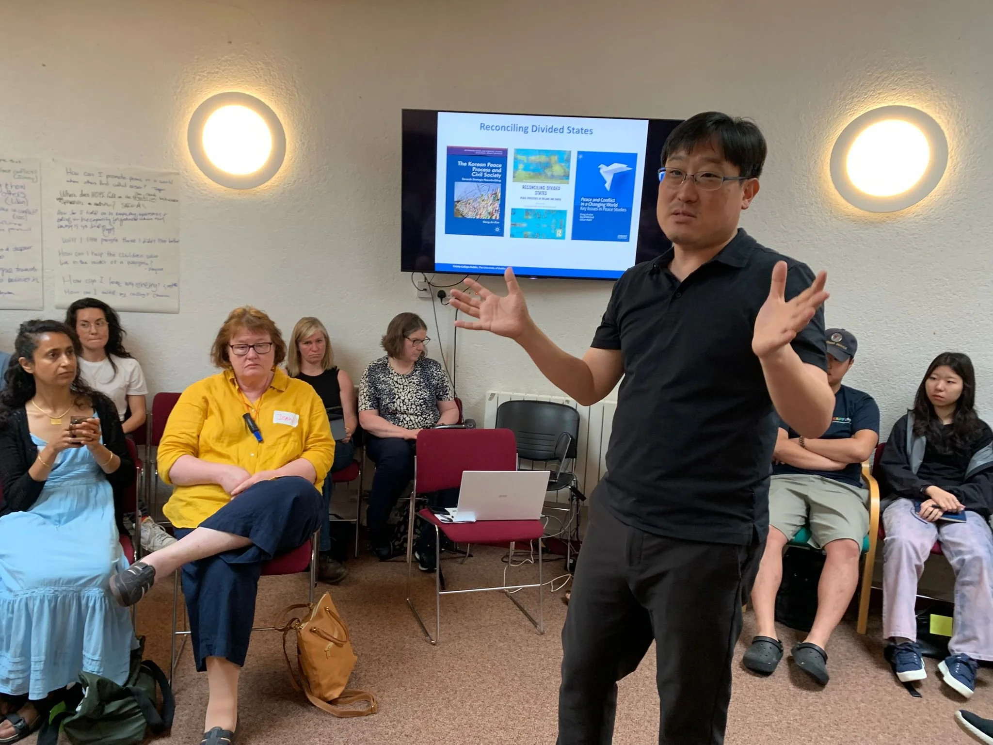 A man in a black shirt and glasses is giving a presentation to a group of people in a room, with a screen behind him displaying slides about reconciling divided states.