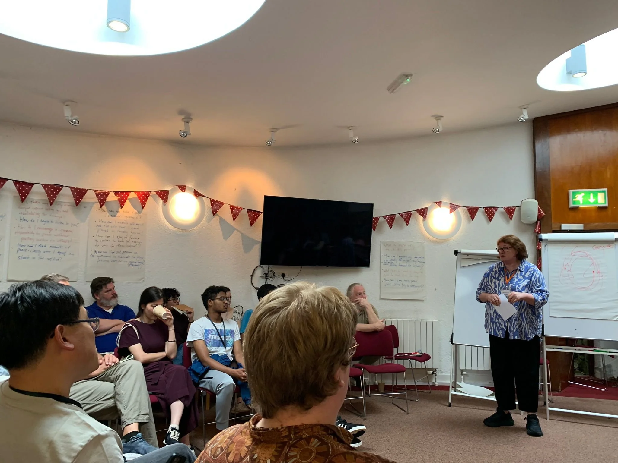 A group of people sitting and listening to a woman standing at the front of a room with a whiteboard and flip chart, decorated with red bunting and circular light fixtures on the wall.