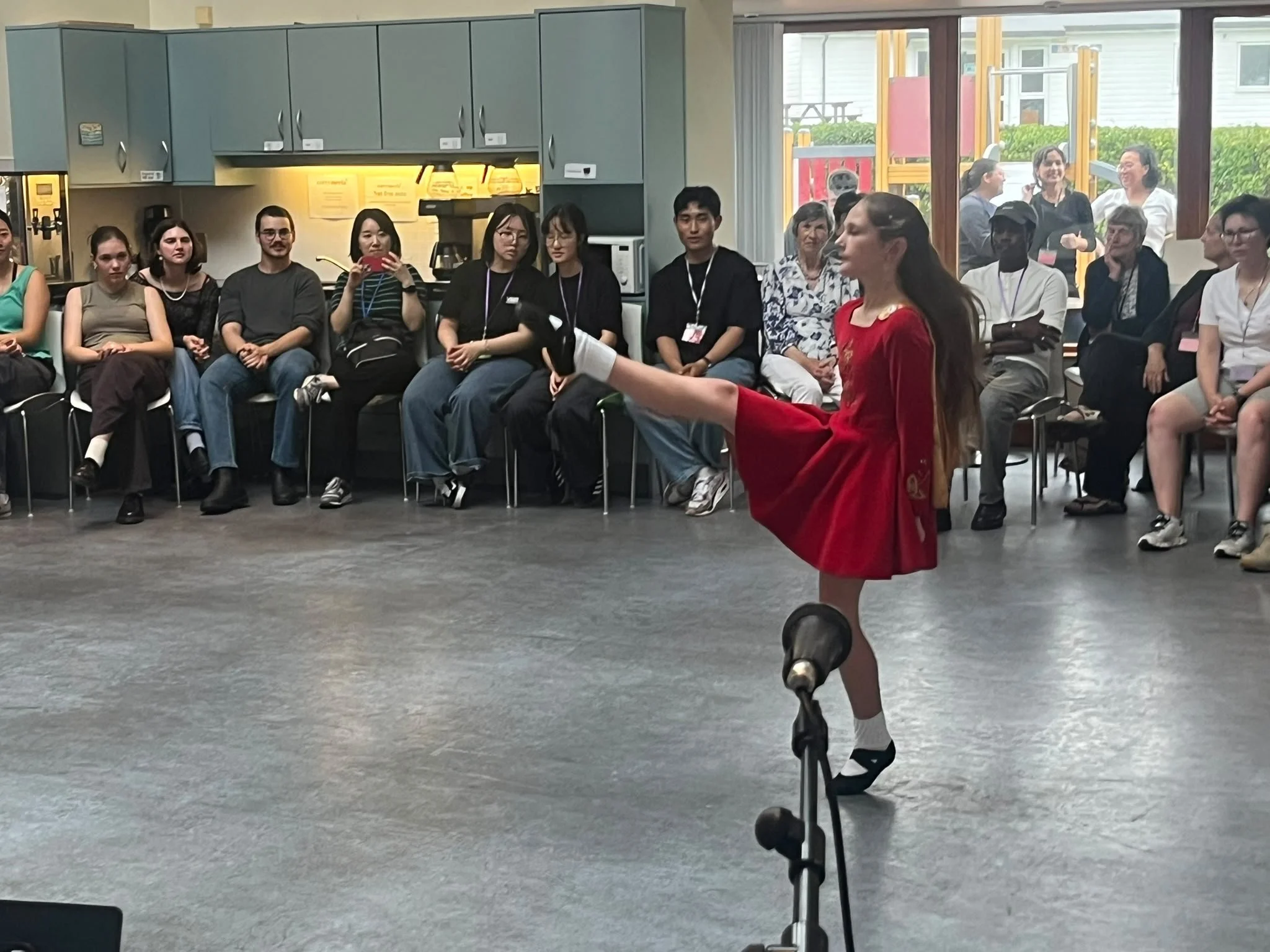 in a red dress and dark hair is performing with a microphone on a stage set up in a room with audience members. Audience members are sitting and watching, some taking photos with their phones. The room has large windows showing an outdoor playground.