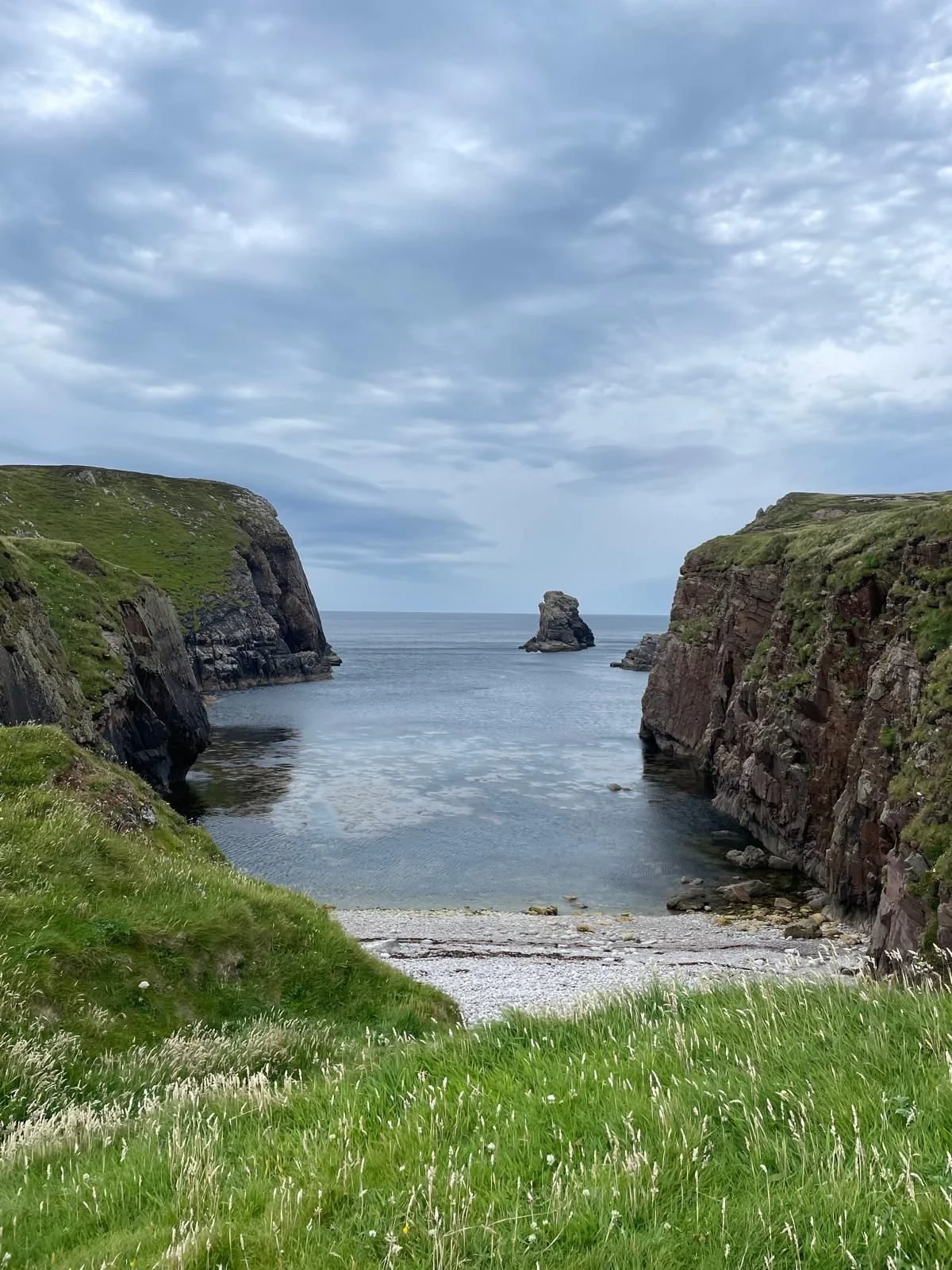 Coastal cove with green grassy cliffs on both sides, a calm water inlet, and a large rock formation in the distance under a cloudy sky.
