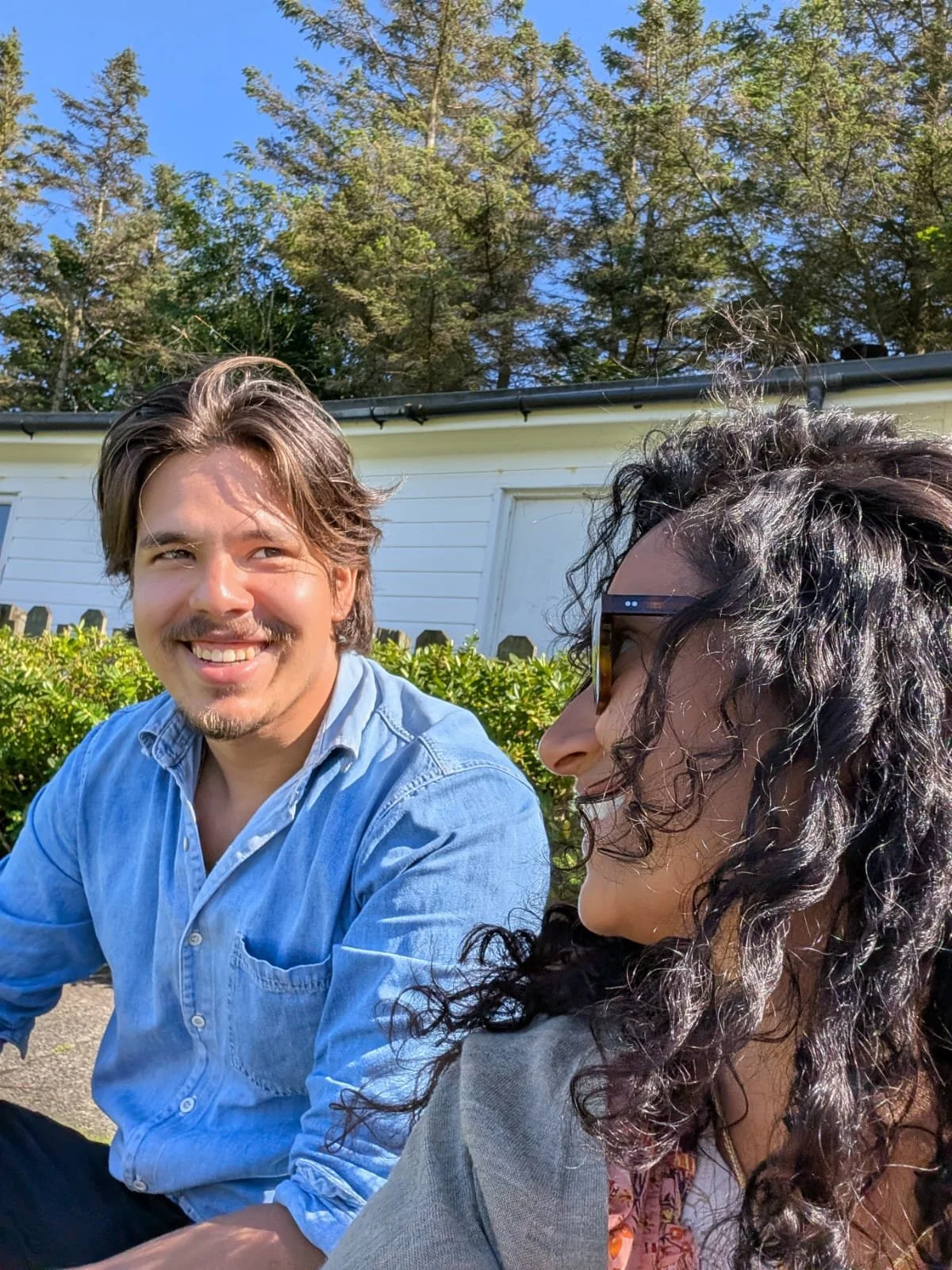 A smiling man with brown hair and a beard wearing a light blue shirt, sitting outdoors next to a woman with curly black hair and glasses, with a white house and trees in the background on a sunny day.