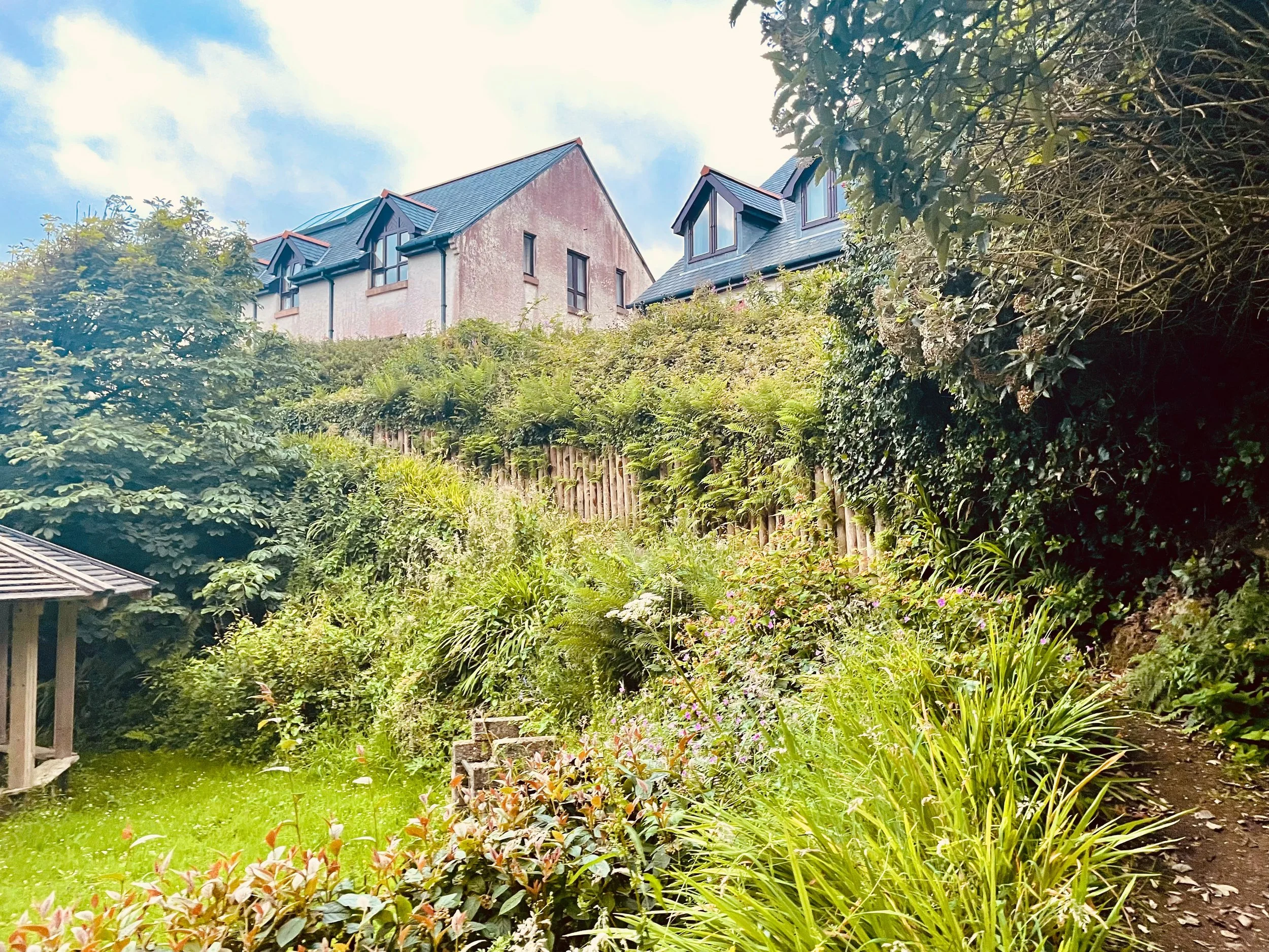 Residential houses behind lush green shrubbery and garden plants, with a partly cloudy sky overhead.