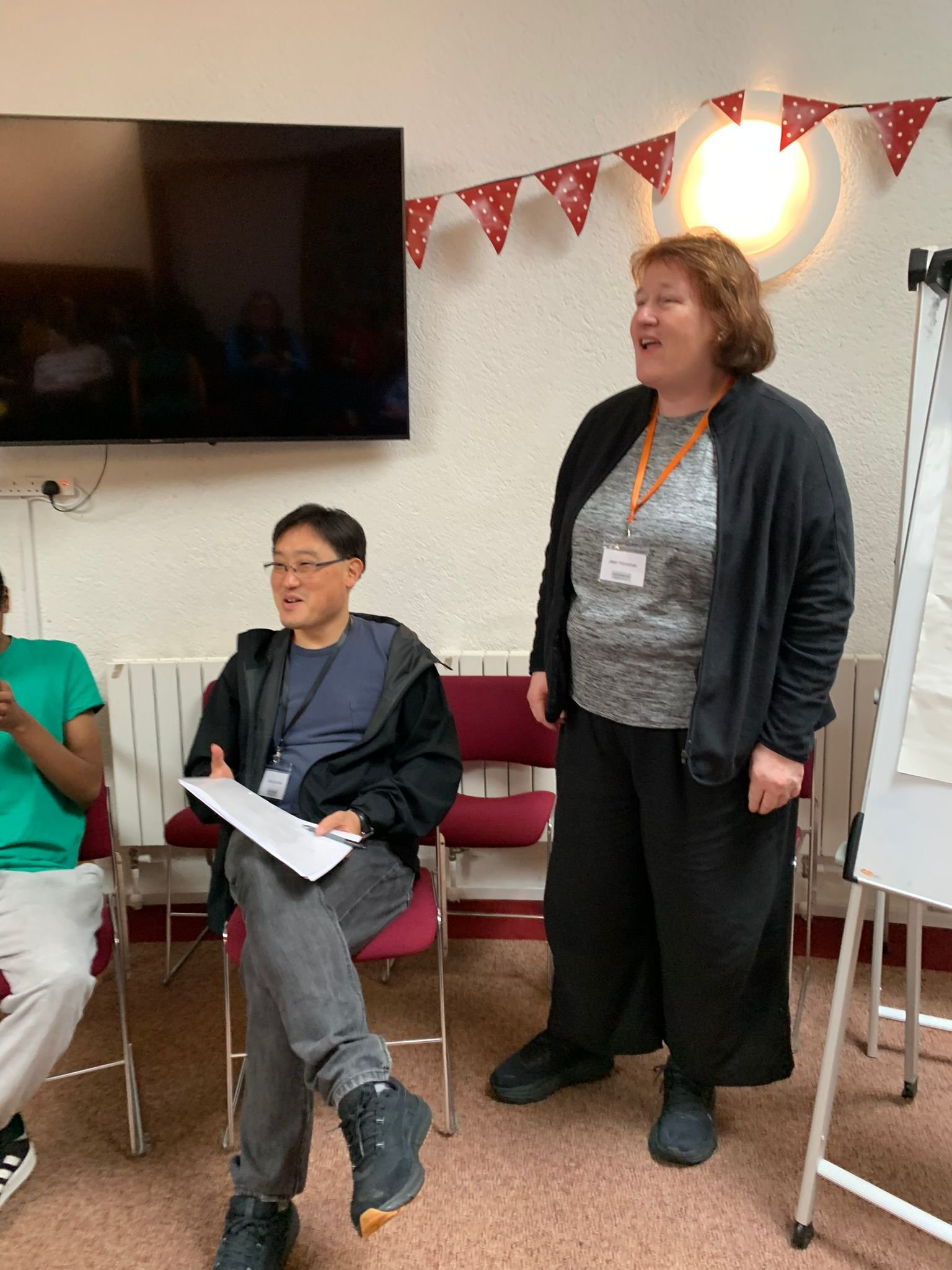 A woman with short, curly red hair standing and speaking, wearing a gray shirt, black jacket, and black pants. A man with glasses, black hair, wearing a gray shirt and black jacket, sitting with a document, and a woman in a green shirt sitting next t