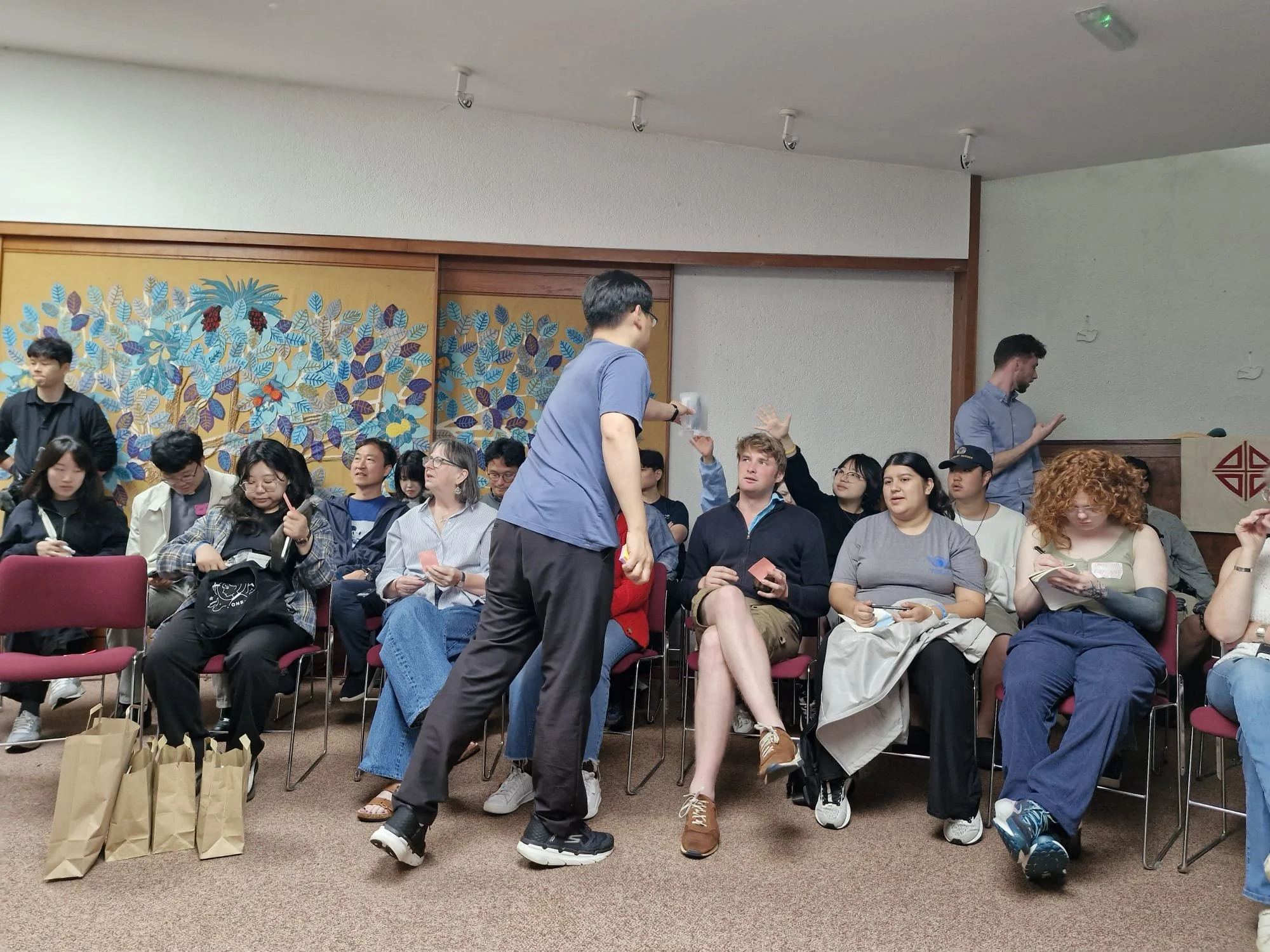People seated in chairs in a room, waiting for an event to start. Some are chatting, taking notes, or holding papers. A person in a blue shirt stands in the center, talking to the seated audience.