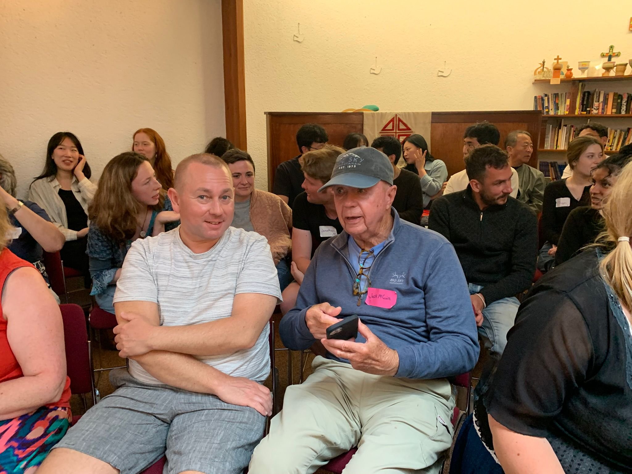 A group of people sitting in a room, engaging in conversation. Two men in the foreground are talking, one with a white striped shirt and the other with a blue jacket and gray cap. The room has a bookshelf and wall decorations.