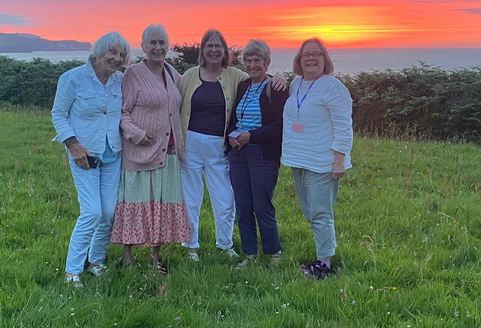 Five women standing together on a grassy field during sunset, with ocean and cliffs in the background, smiling at the camera.