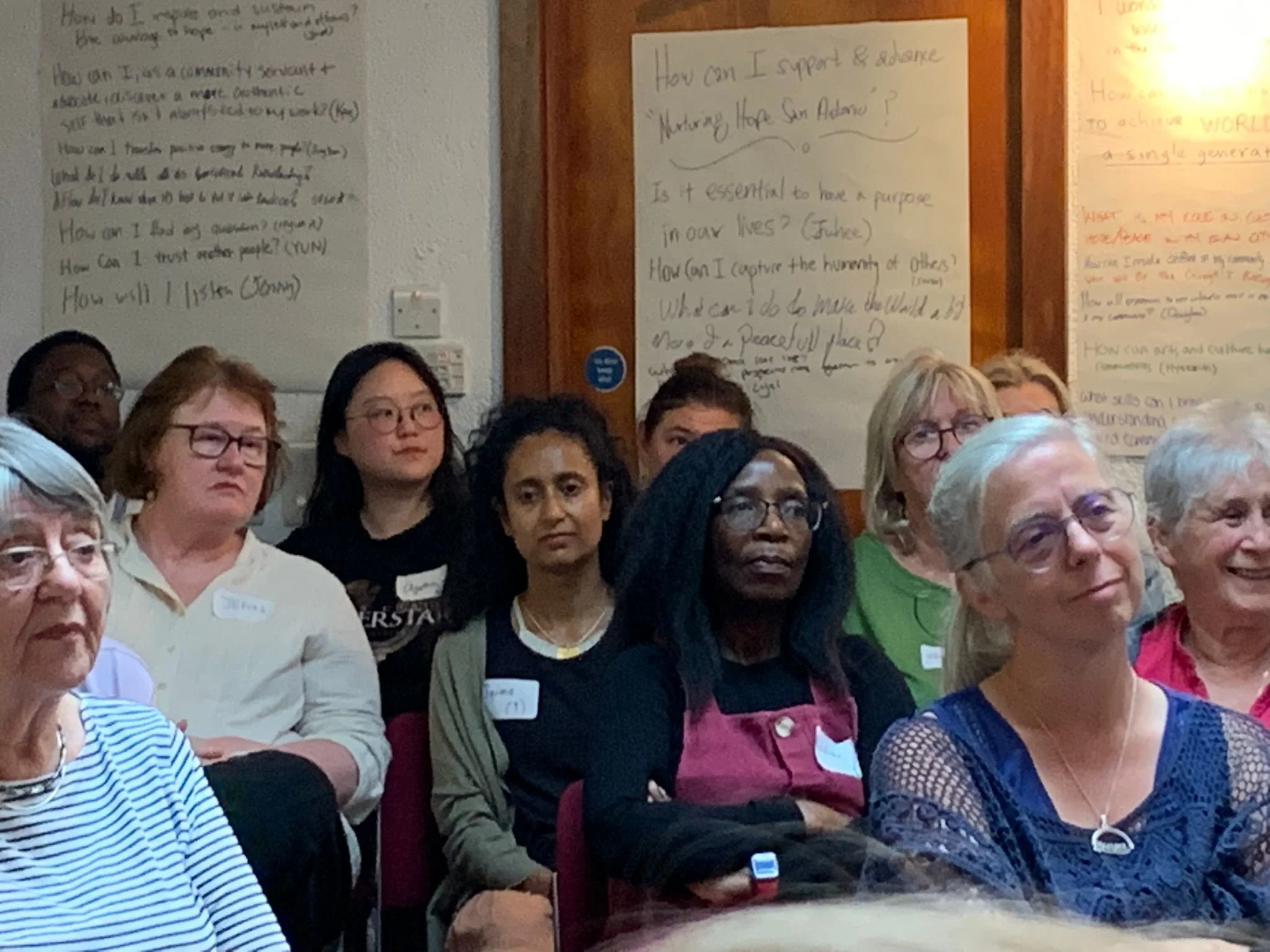 A group of diverse women attending a workshop or seminar, sitting and listening attentively. There are handwritten posters with questions on whiteboards or large sheets in the background.