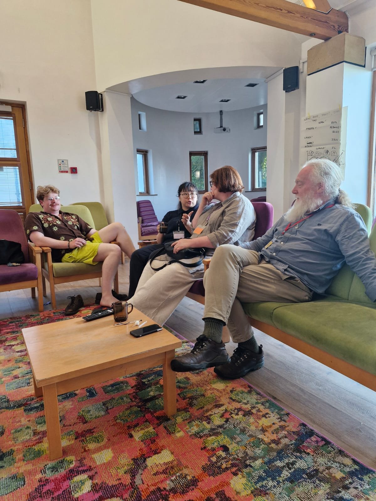 Four people sitting and talking in a cozy, well-lit indoor space with colorful chairs, a small wooden table, and a patterned rug.