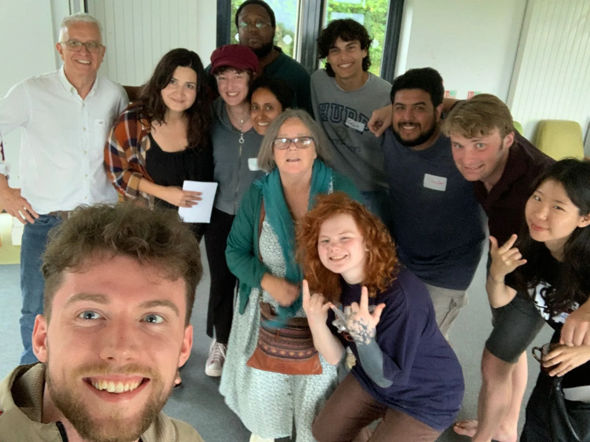 Group of diverse young adults and older individuals smiling and posing for a photo indoors, some making hand gestures, in a well-lit room with windows.