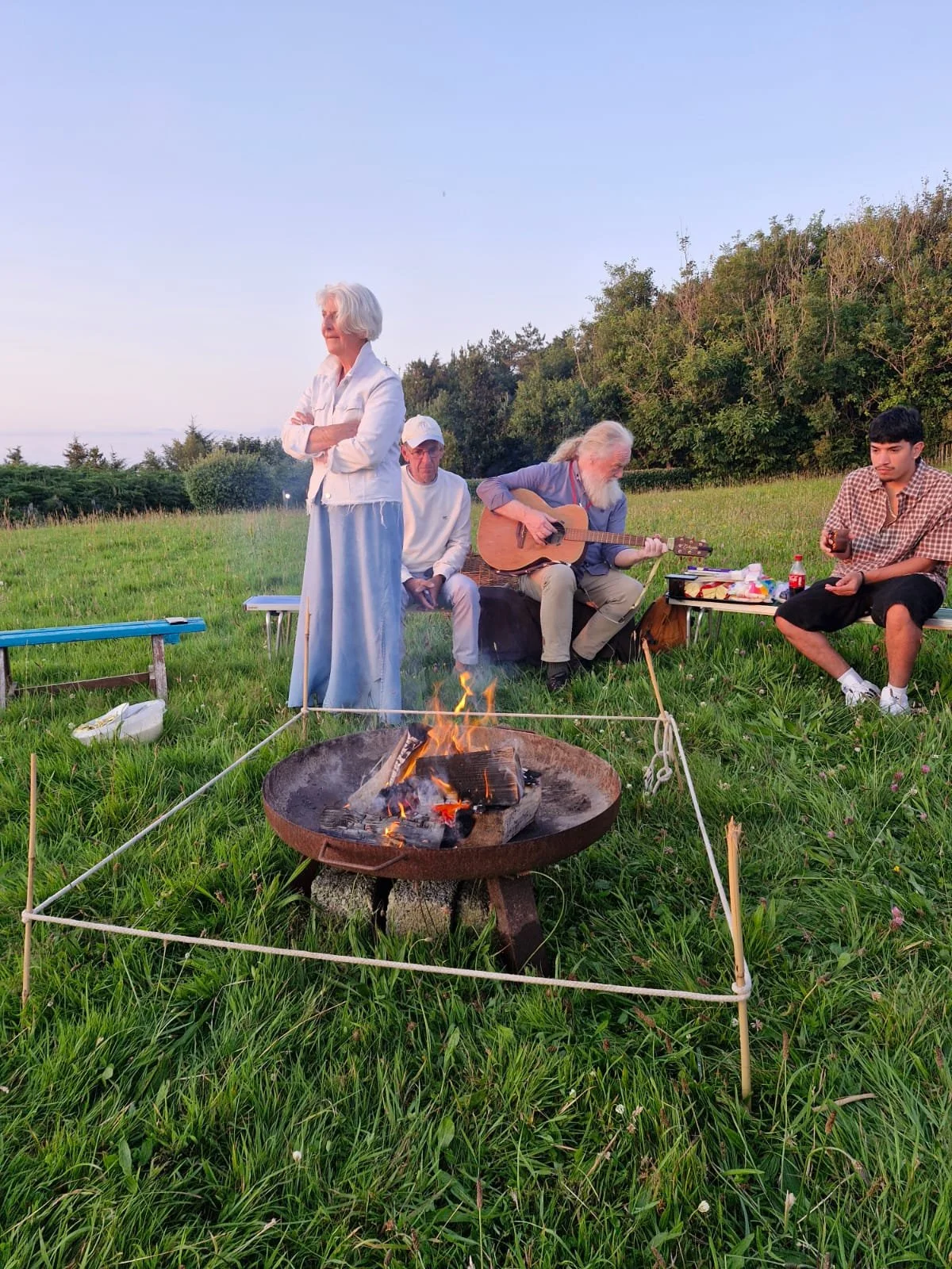 People enjoying an outdoor gathering around a campfire, with one person playing guitar, another with crossed arms, and others seated on benches