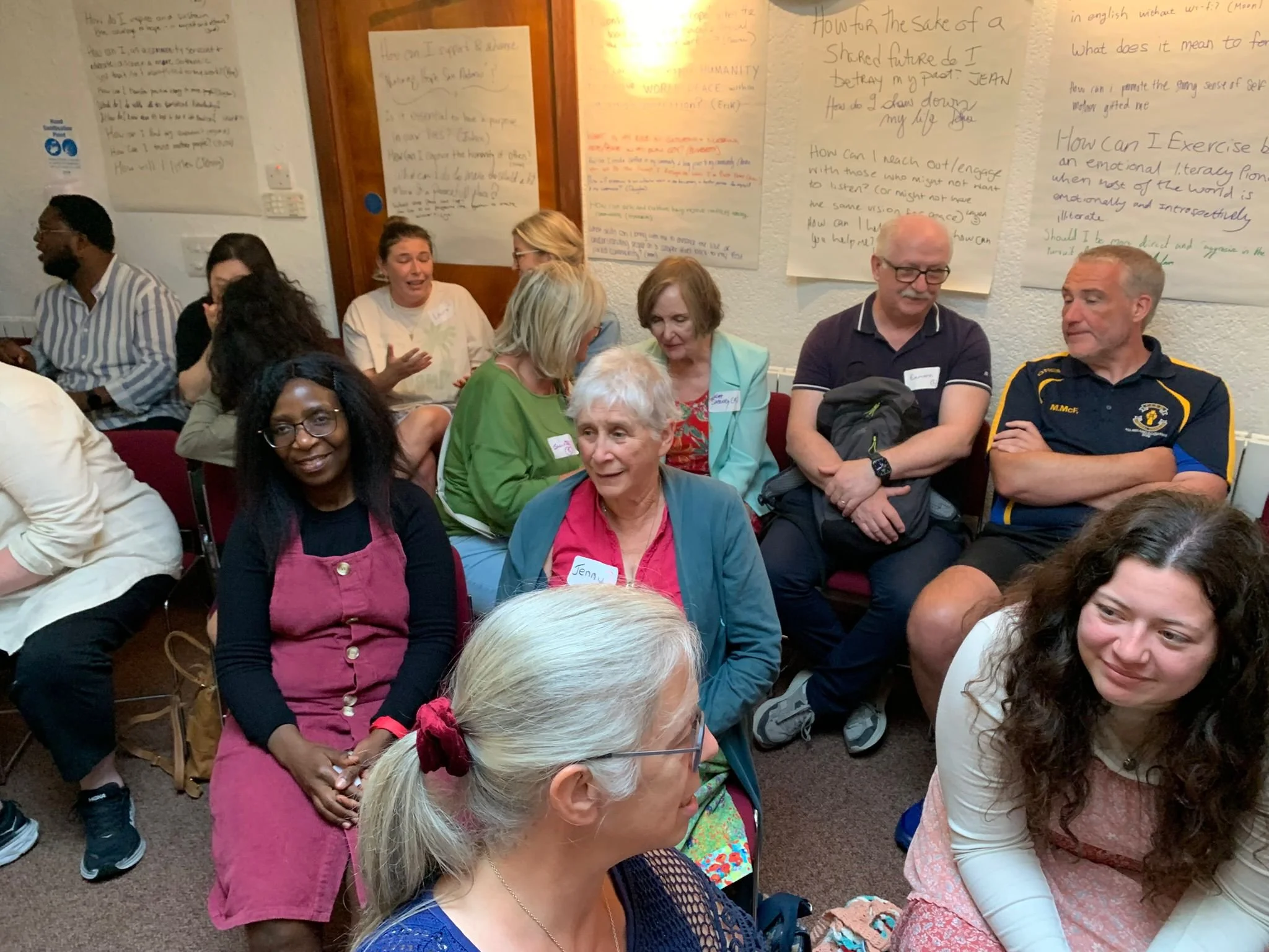 Group of diverse adults seated in a room, engaging and listening during a workshop or seminar, with handwritten posters on the walls.