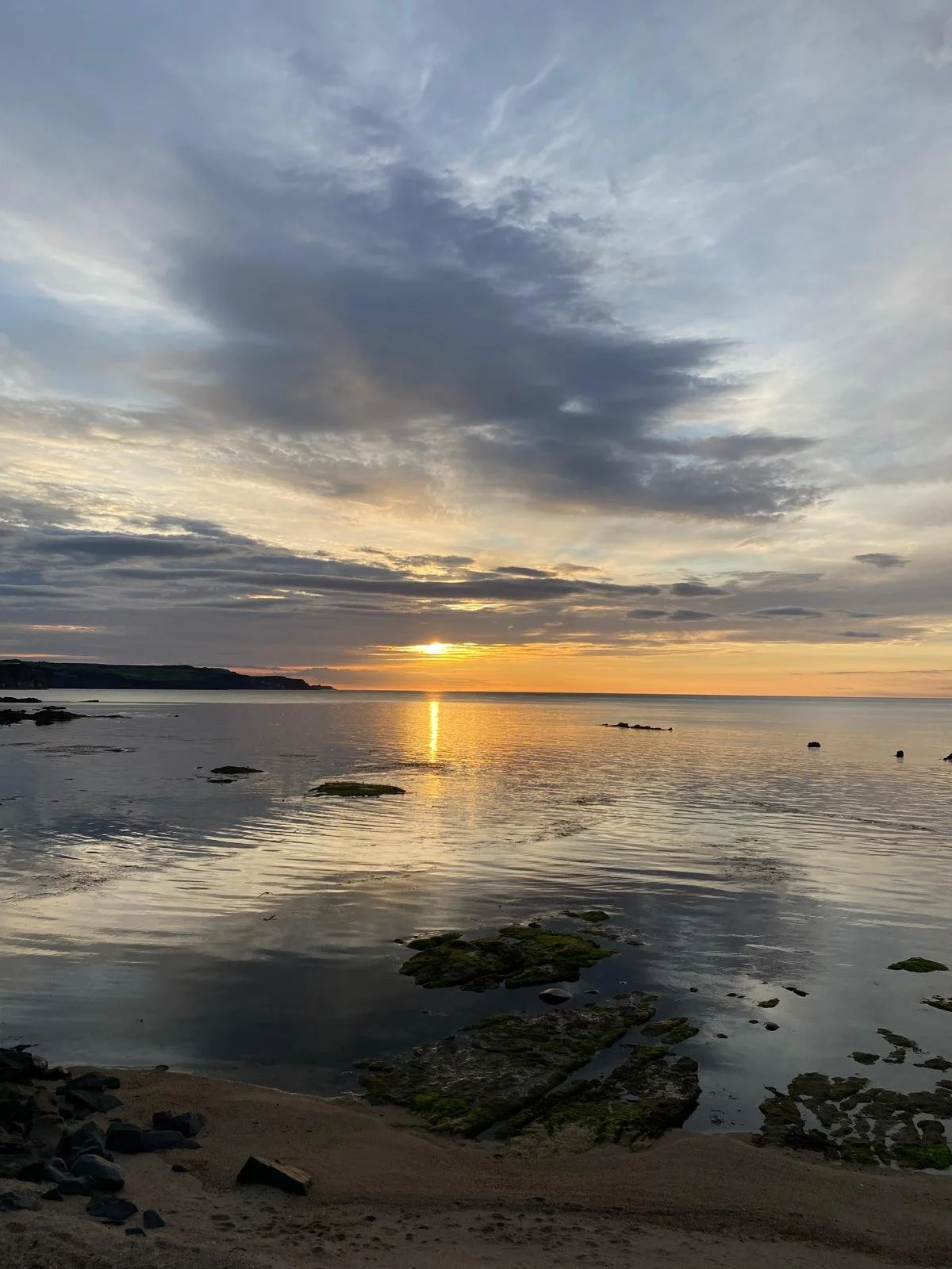 Sunset over a calm ocean with light clouds and rocks on the shore.