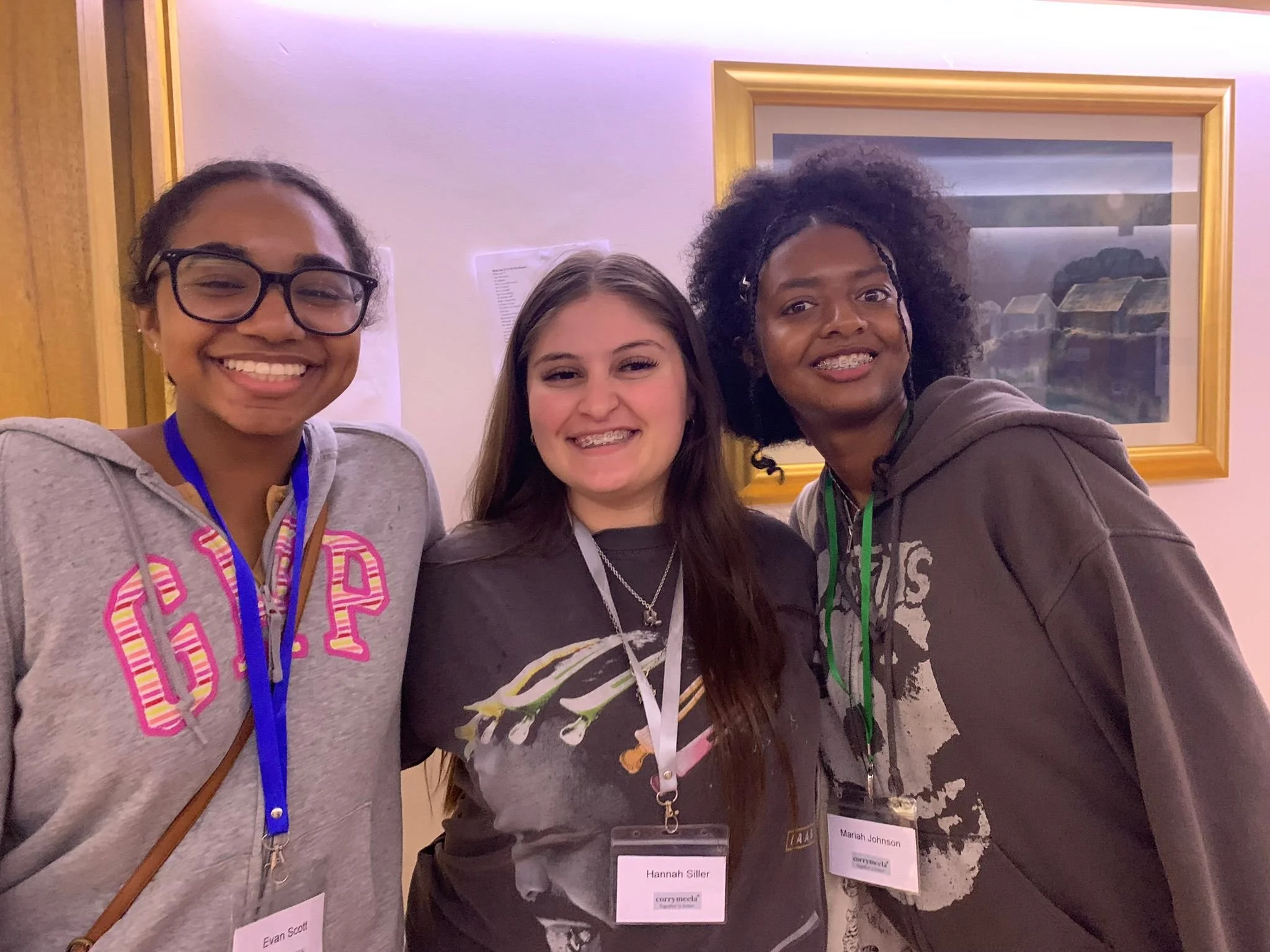 Three young women smiling and standing close together indoors, wearing conference badges around their necks.