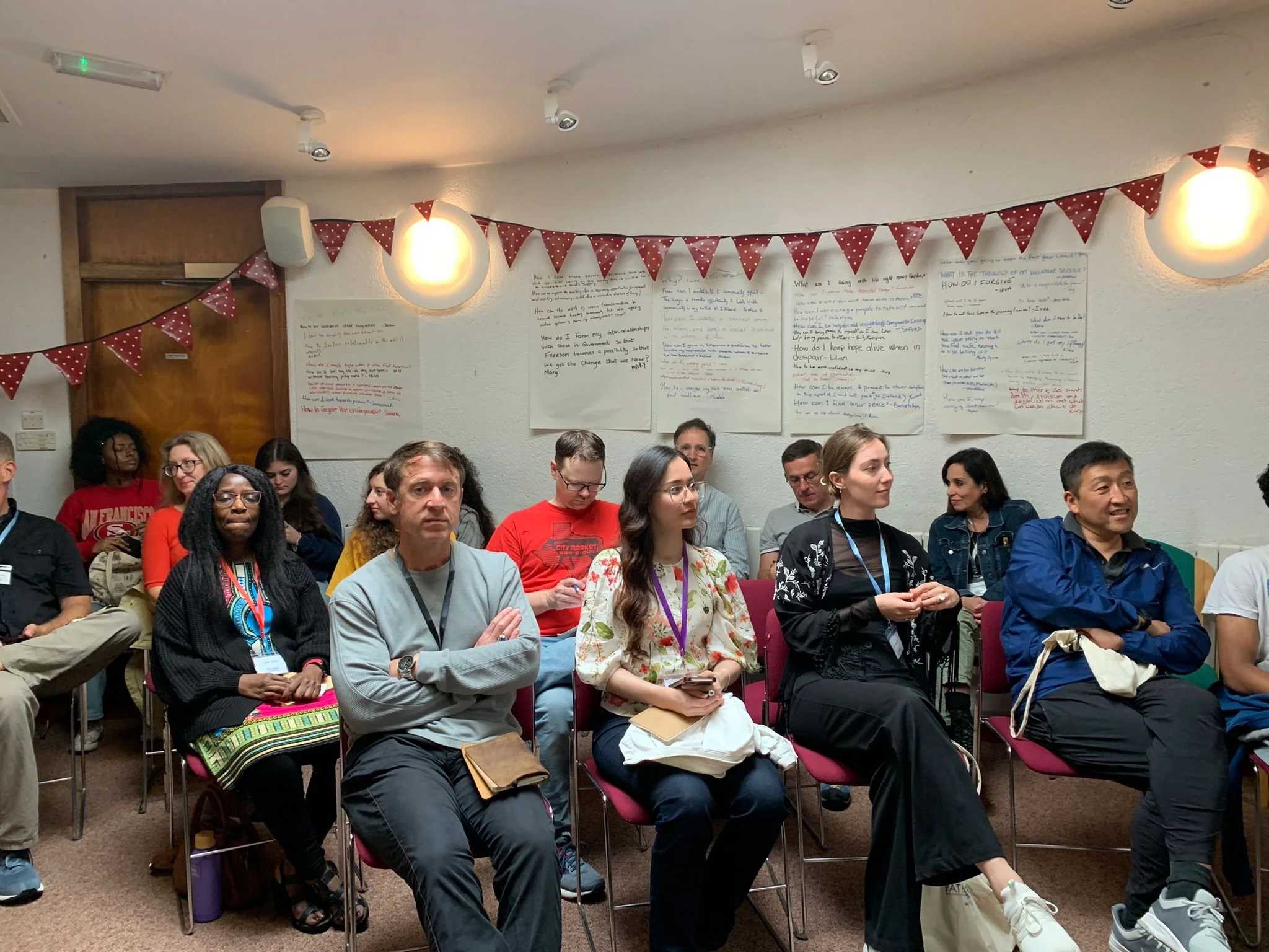 People seated in a conference room attending a presentation or workshop, with handwritten posters on the wall and red bunting decorations.