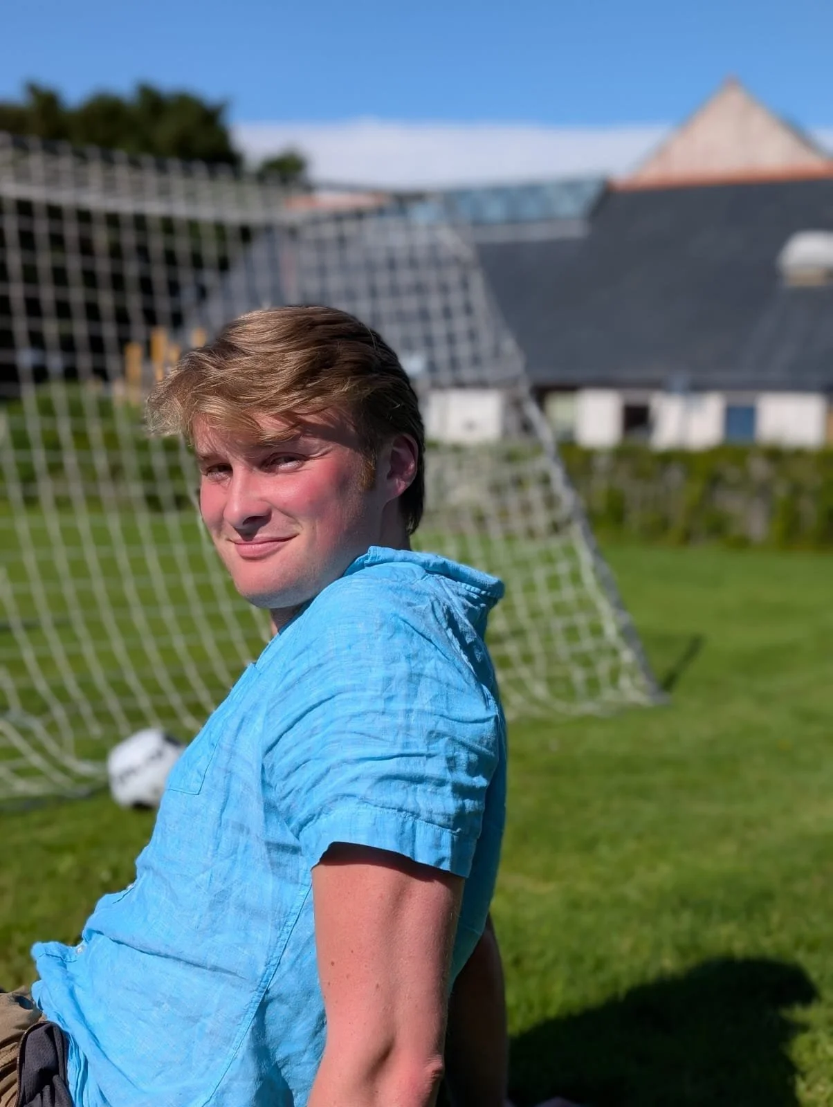 Young man with light brown hair smiling outdoors on a sunny day, wearing a light blue shirt, in front of a soccer goal net with a soccer ball on the grass.