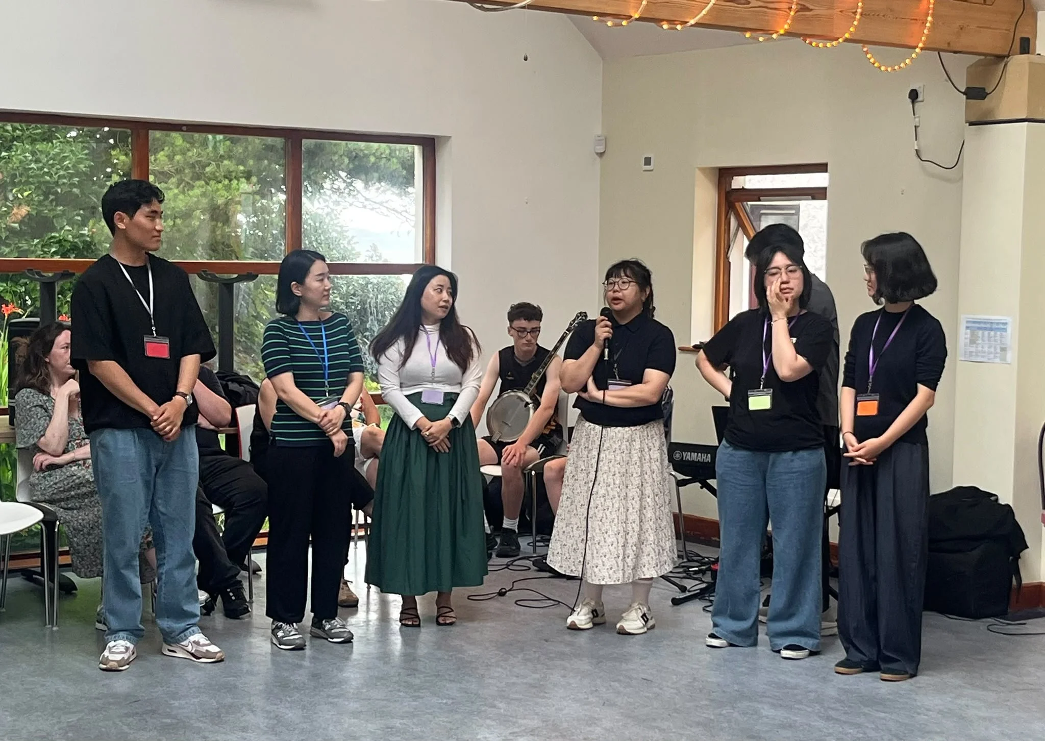 A group of seven people standing in a semi-circle, some holding microphones, in a room with large windows and a wooden ceiling with string lights. Others are seated in the background, one with a guitar, and a small keyboard is seen on the right.