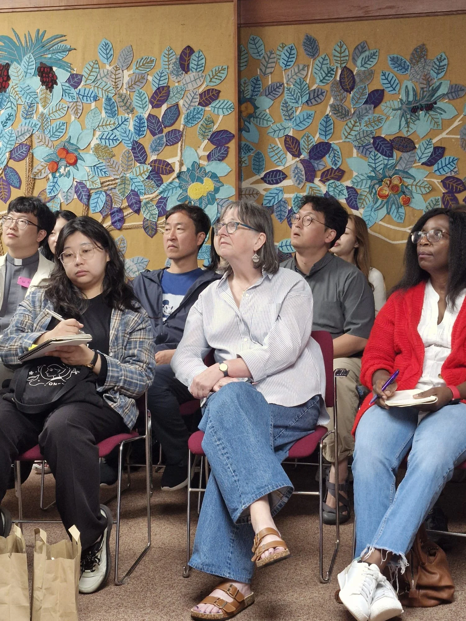 People attentively listening to a presentation in a room with a decorative wall featuring colorful leaves and berries.