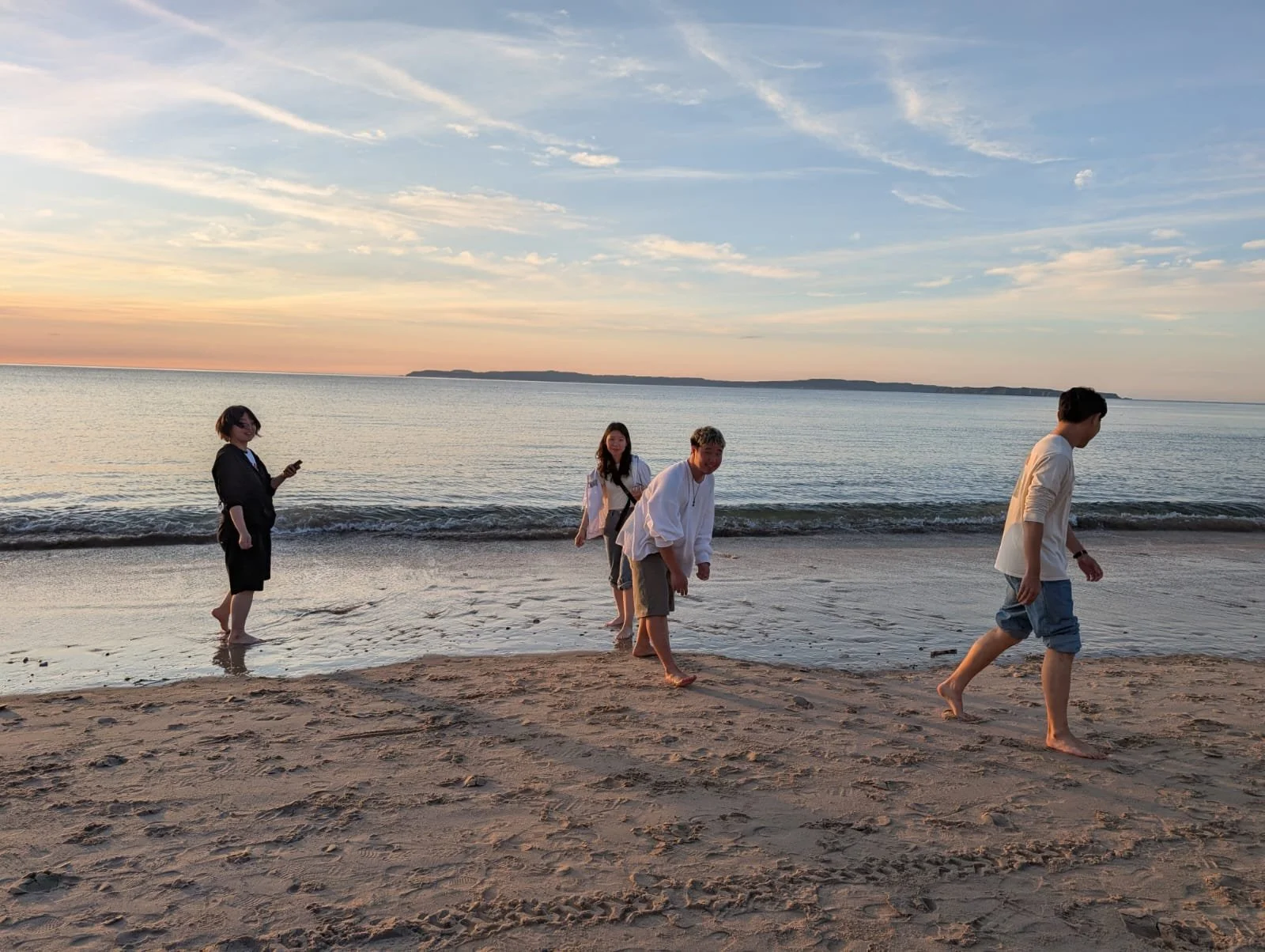 Four people walking on a sandy beach by the ocean during sunset, with a distant landmass on the horizon, some of them barefoot and one person looking at their phone.