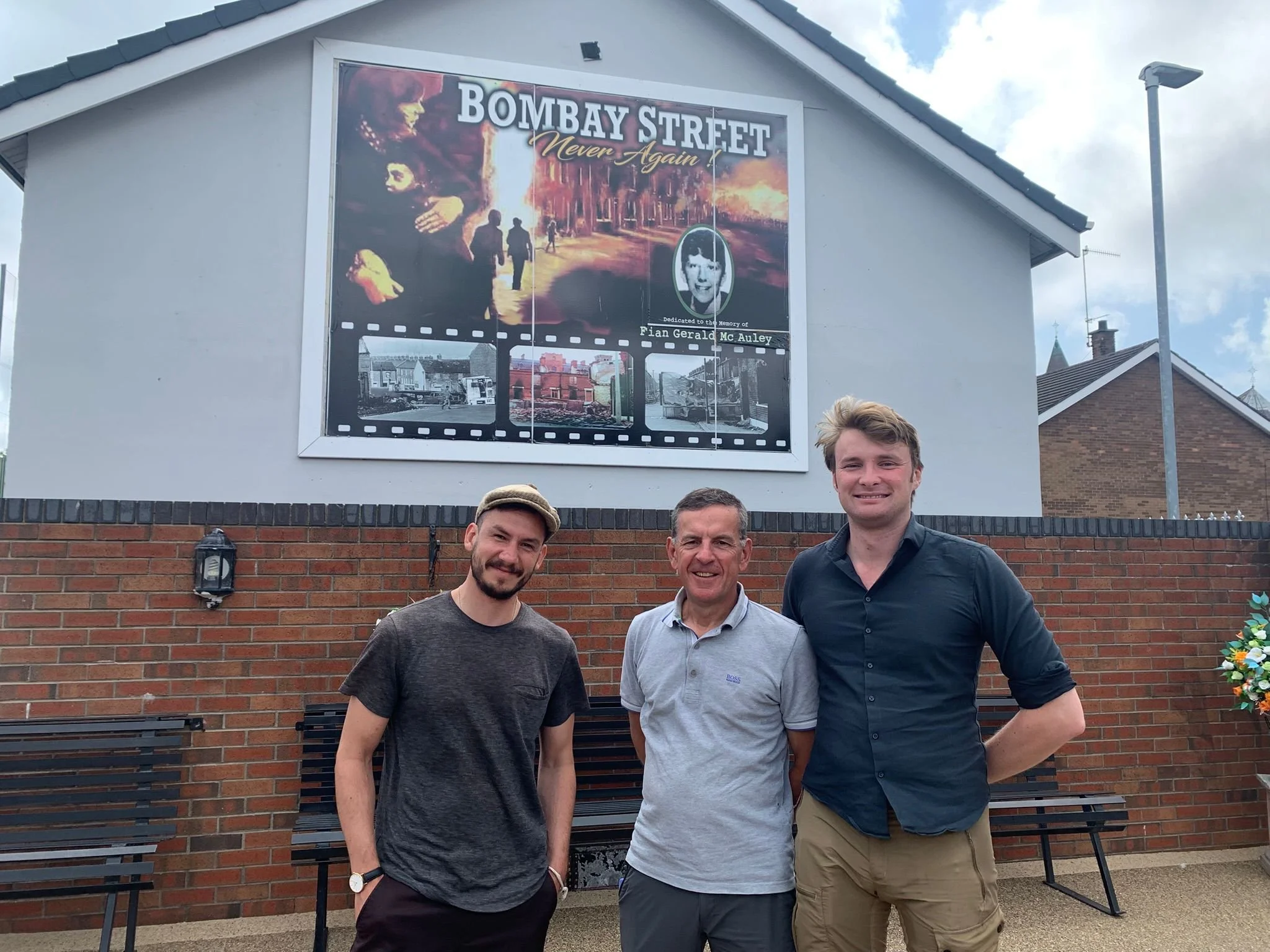 Three men standing outdoors in front of a brick wall with a large billboard displaying a poster for the film 'Bombay Street Never Again'.
