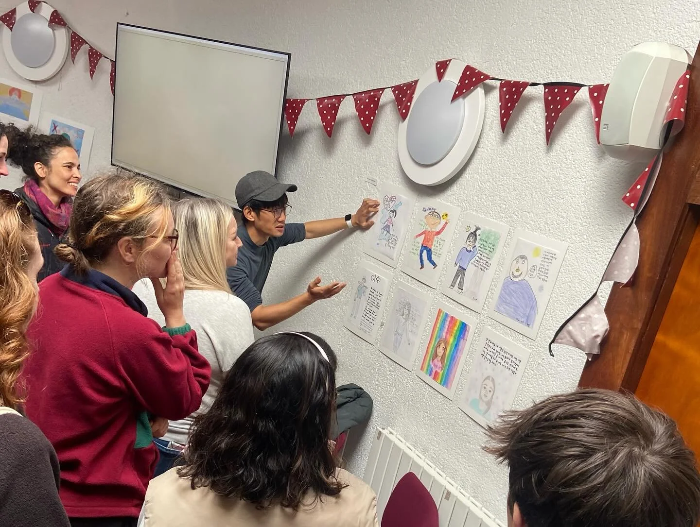 A group of people listening to a man presenting a display of colorful drawings and writings on a wall, decorated with red polka dot bunting.
