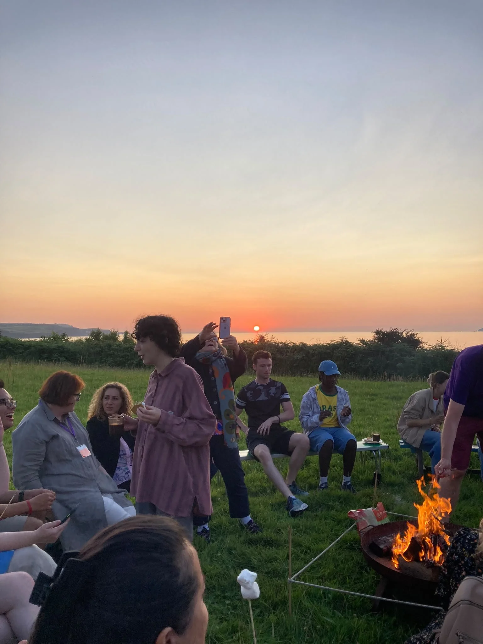 A group of people gathered outdoors around a small fire at sunset, with green grass and trees in the background.