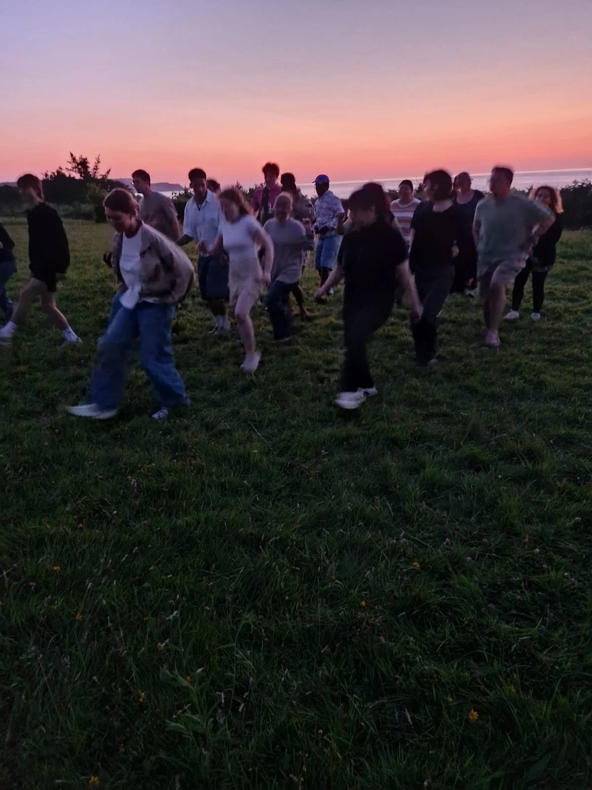 People gathered outdoors during sunset, walking or dancing on grass with a view of the ocean in the background.