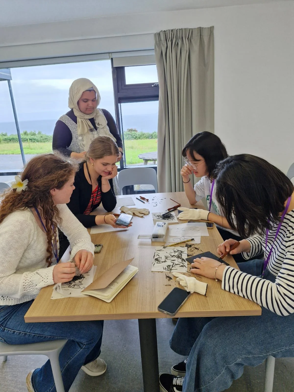 Group of women working on printmaking at a table, examining and discussing them, with some taking notes or using a phone.