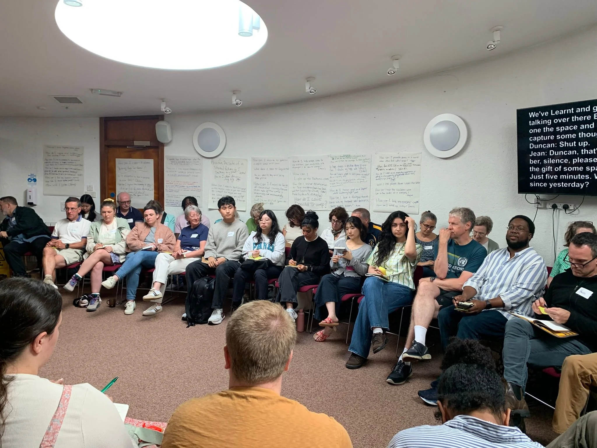 A group of diverse people sitting on chairs in a conference room, facing forward. There are whiteboards with handwritten notes on the wall, and a large screen displaying text on the right side of the image.