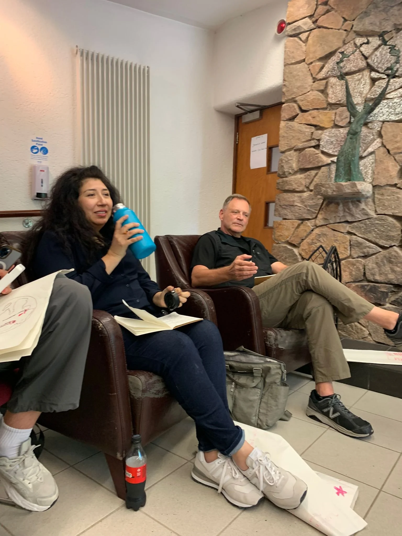 People sitting on a brown leather sofa in a waiting area, with one woman holding a blue water bottle, another man with a notepad, and someone partially visible holding a phone. There is a stone wall with a metal sculpture of a tree and a large wooden