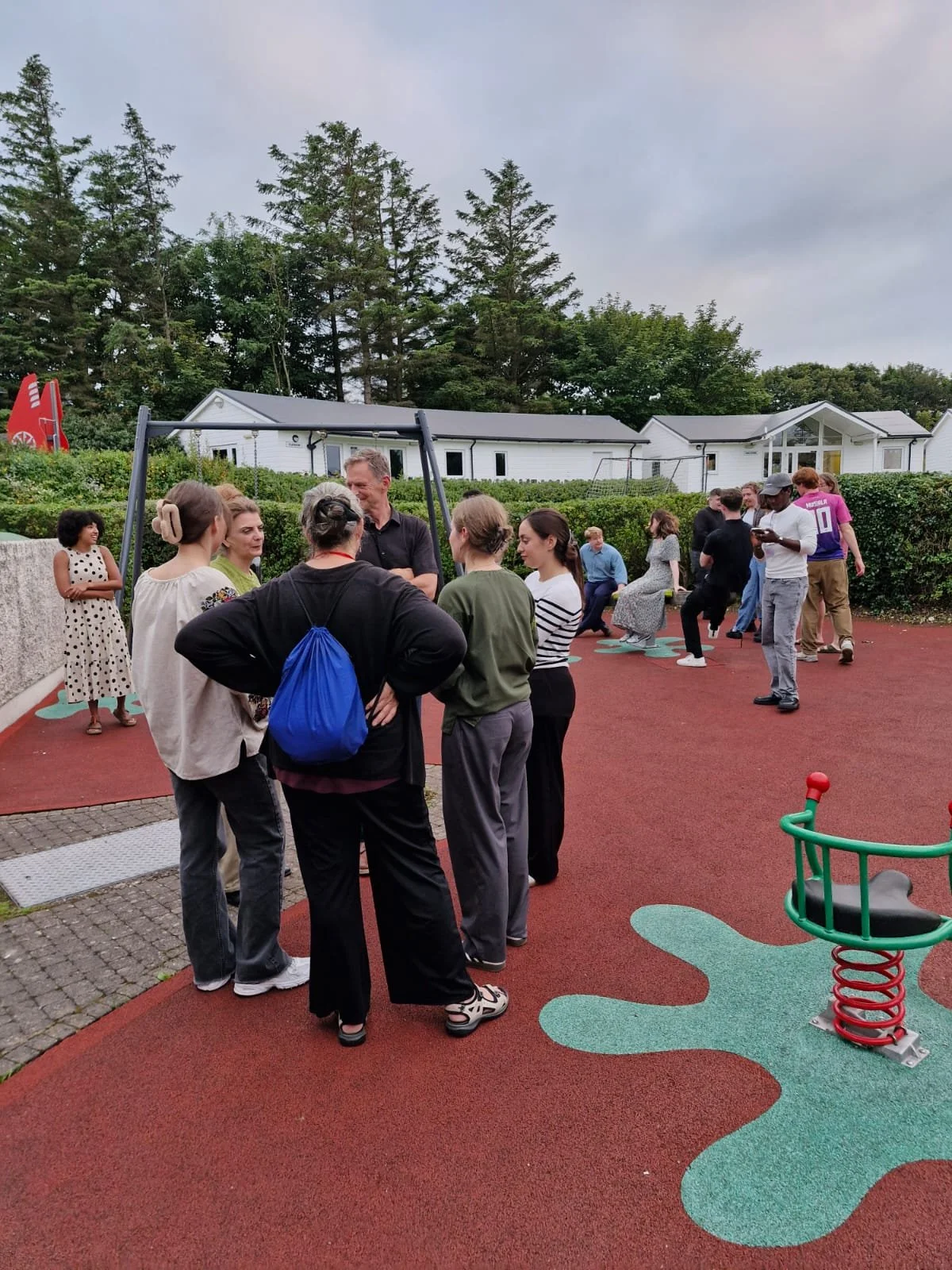 Group of people socializing and playing on a playground with a red rubber surface, surrounded by trees and white buildings.
