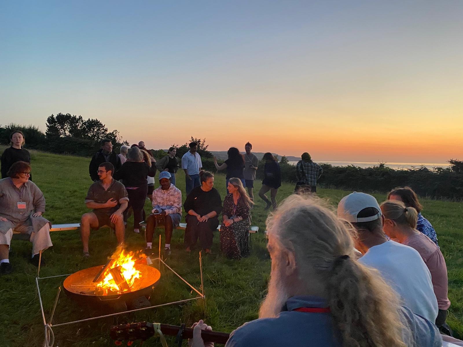 People gathered around a campfire on a grassy hill at sunset with trees and water in the background.