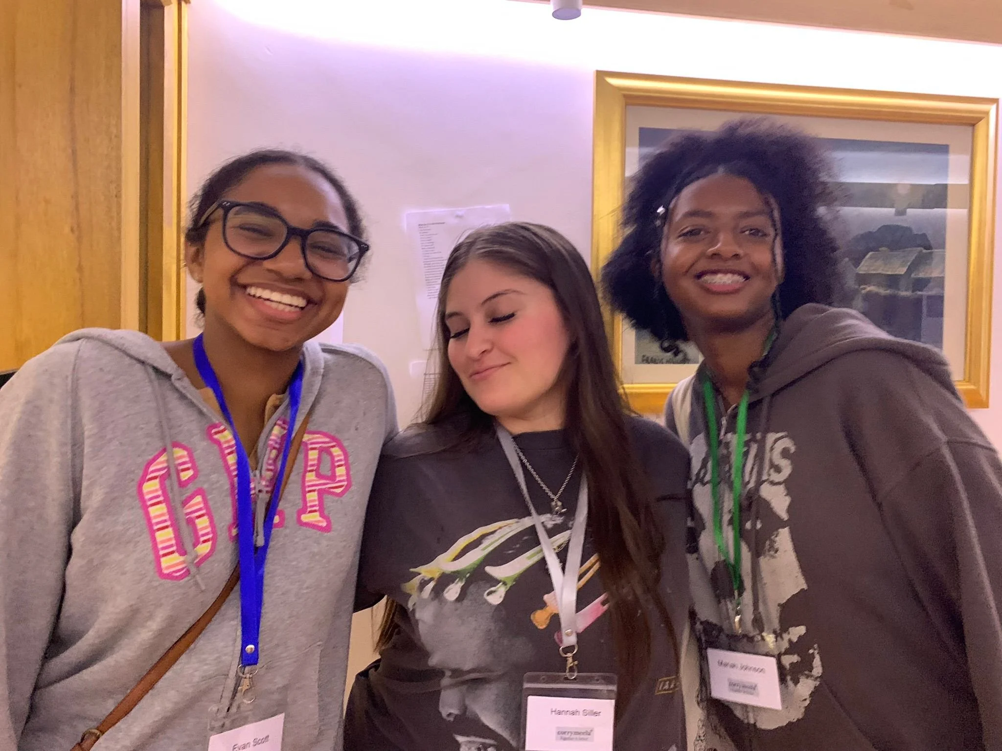 Three young women posing together at an indoor event. The woman on the left is smiling, wearing glasses, a grey hoodie with pink lettering, and a blue lanyard. The woman in the middle is looking down slightly, with long hair, wearing a black shirt wi