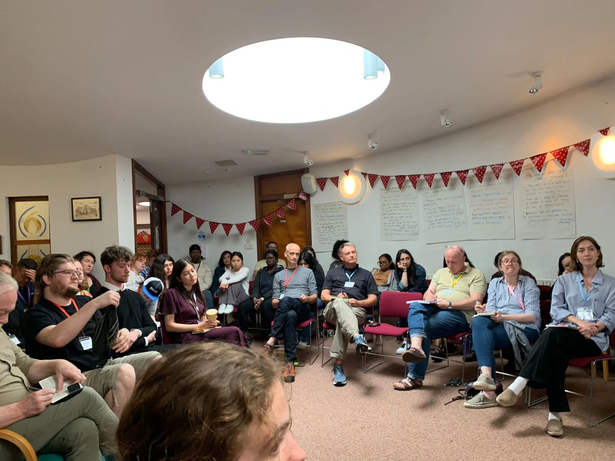 A diverse group of people sitting in a circle in a conference room during a meeting or workshop, with some taking notes and others listening attentively, red and white bunting hanging on the wall, whiteboard with handwritten notes, and a large ceilin