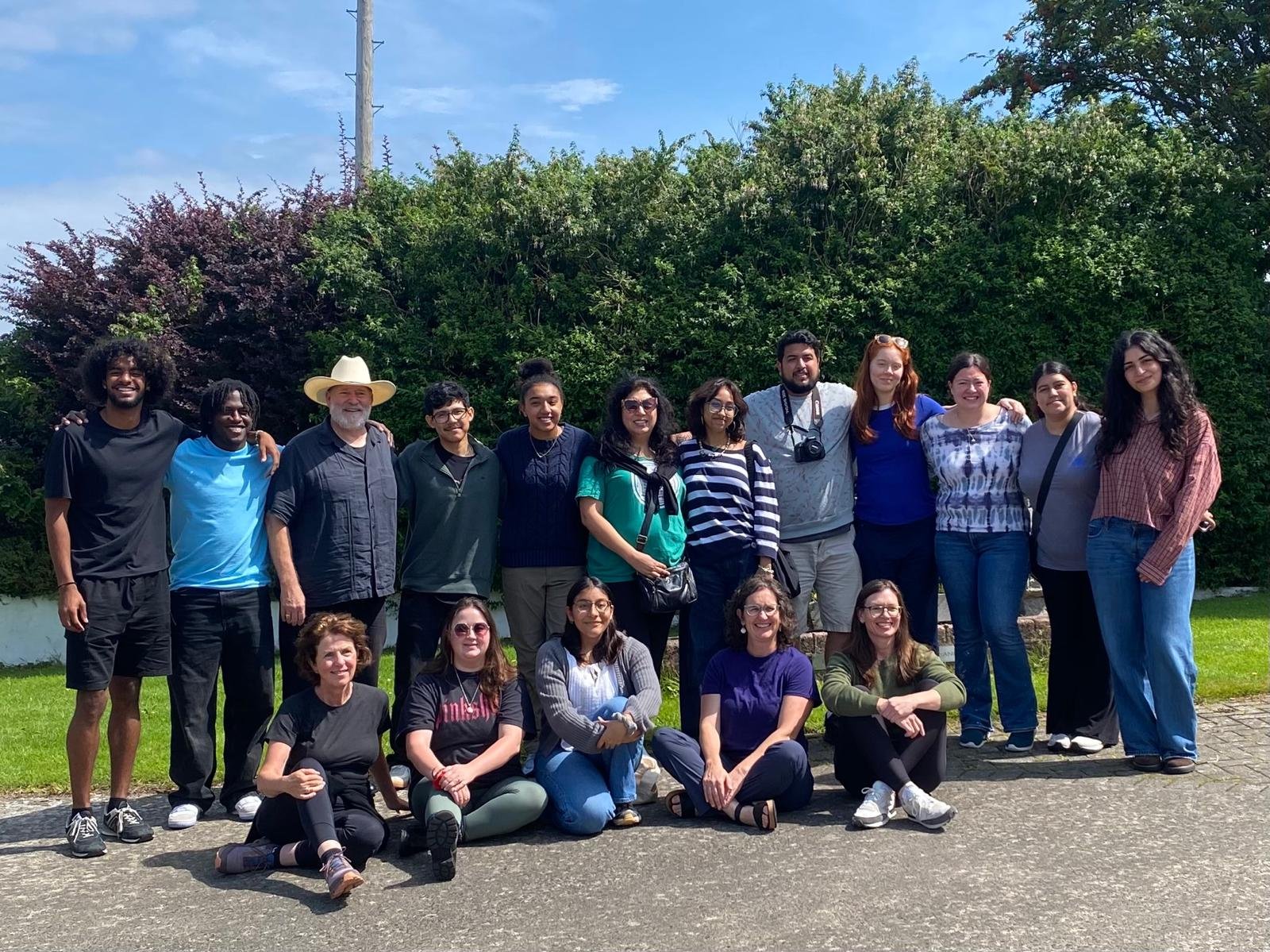 Group of 15 diverse people standing and sitting outdoors, smiling, in front of green bushes and blue sky.