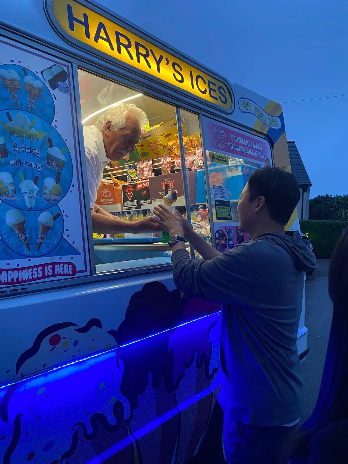 A young man is reaching out to an older man in an ice cream truck named 'Harry's Ices', with the truck illuminated in blue lights in the evening.