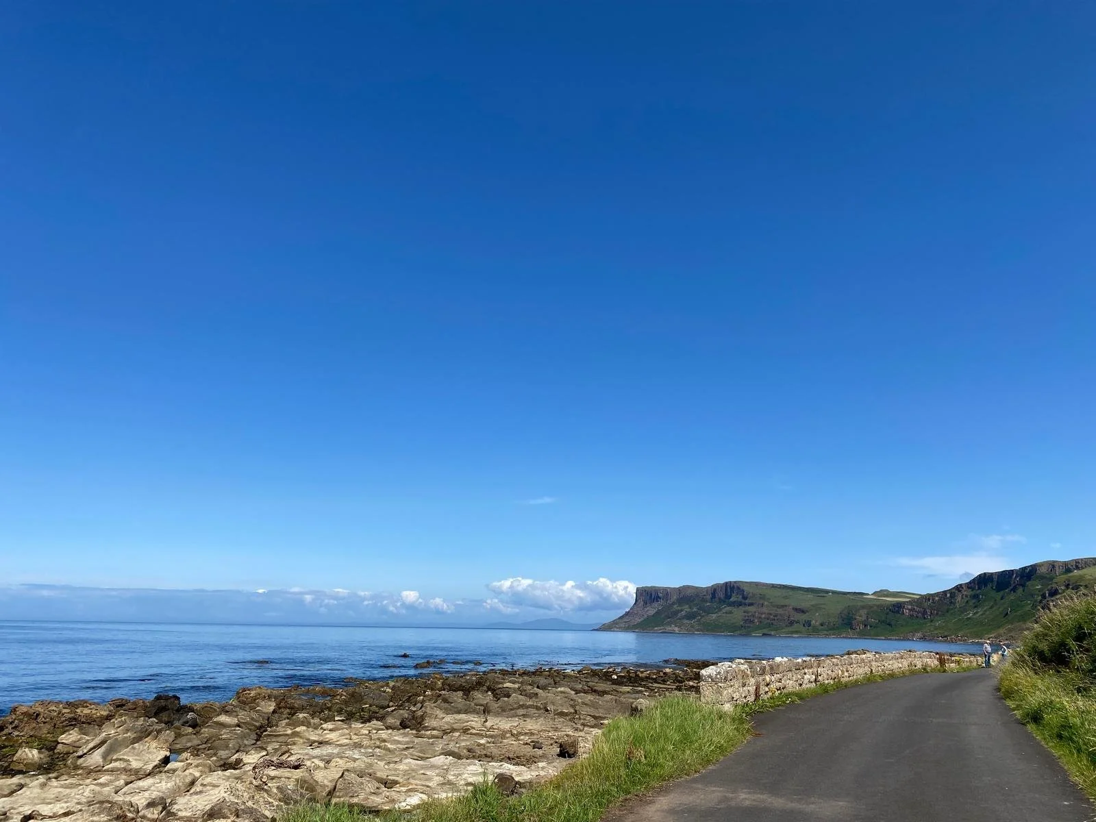 A coastal landscape with a rocky shoreline and a narrow paved road running alongside the ocean under a clear blue sky.