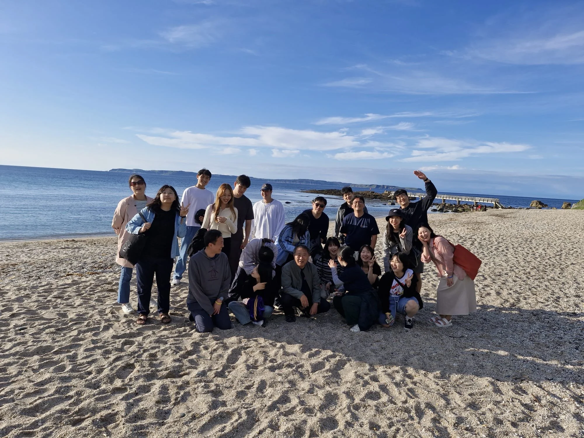 A group of people on a sandy beach with the ocean and a pier in the background, enjoying a sunny day.