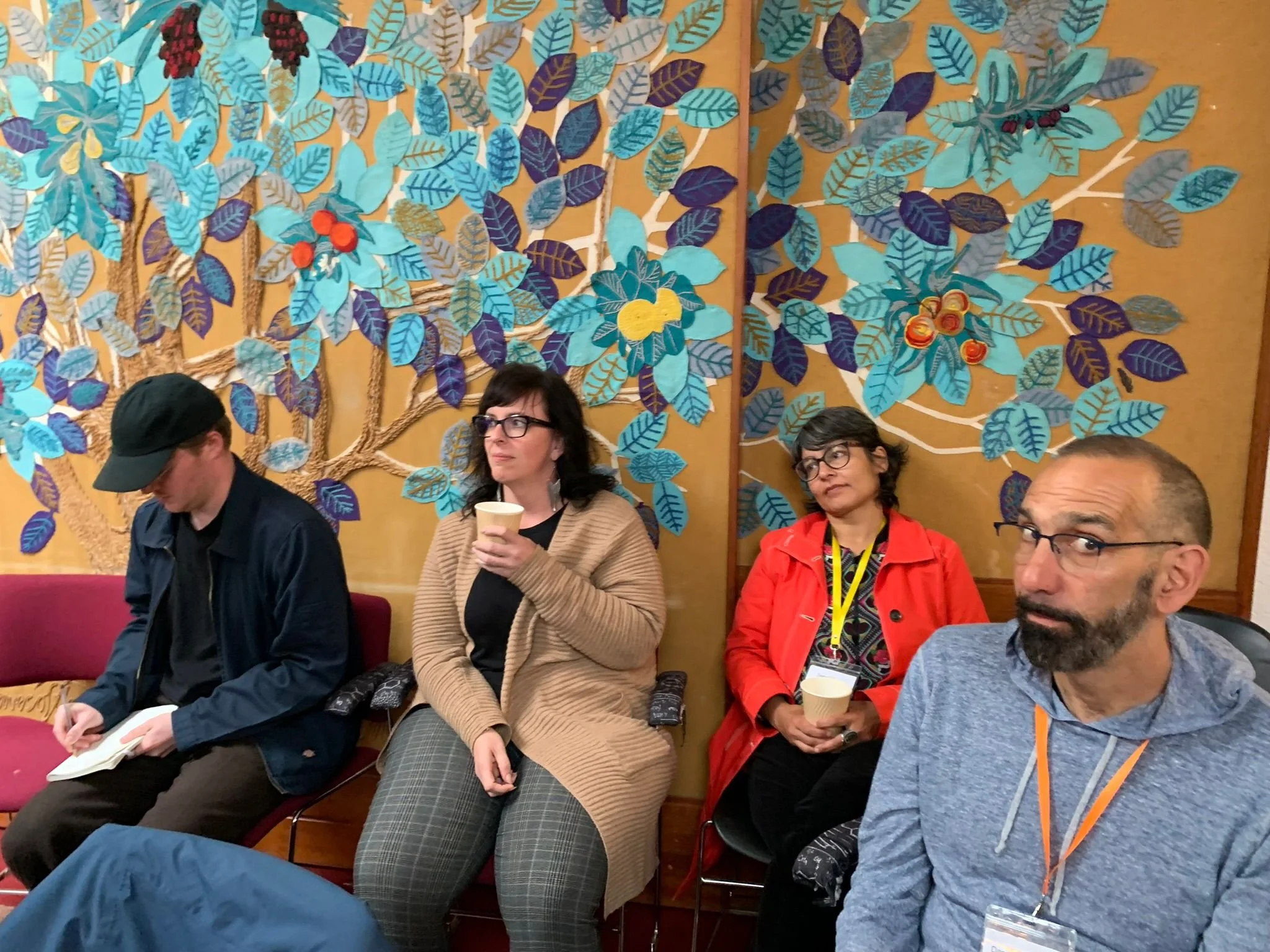 Four people sitting against a decorative wall with blue and purple leaves and fruit patterns, each holding a cup or notepad, with serious and contemplative expressions.
