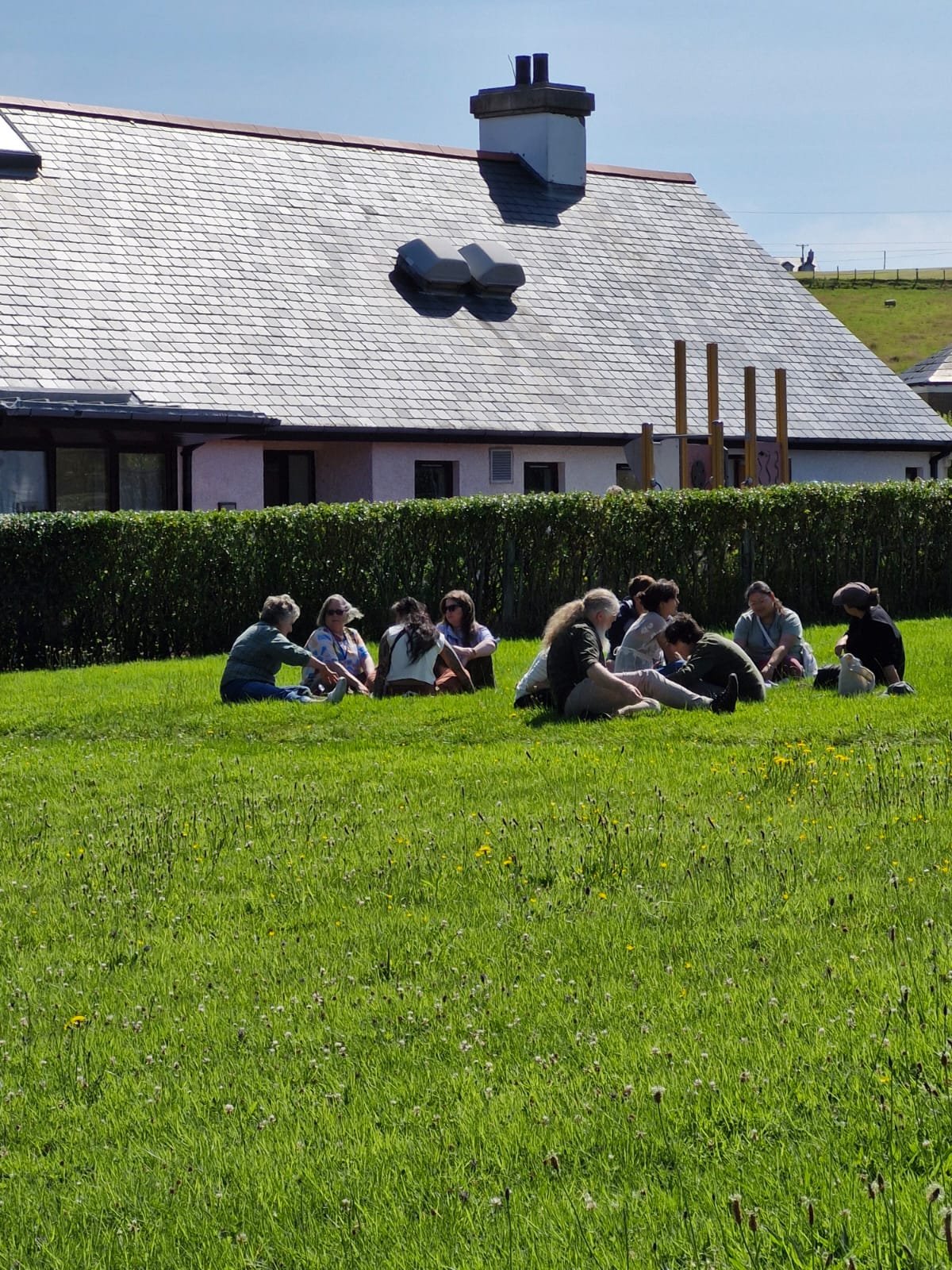 Group of people sitting on a grassy field in a park, talking and enjoying the outdoors on a sunny day. In the background, there is a house with a sloped roof and a hedge.