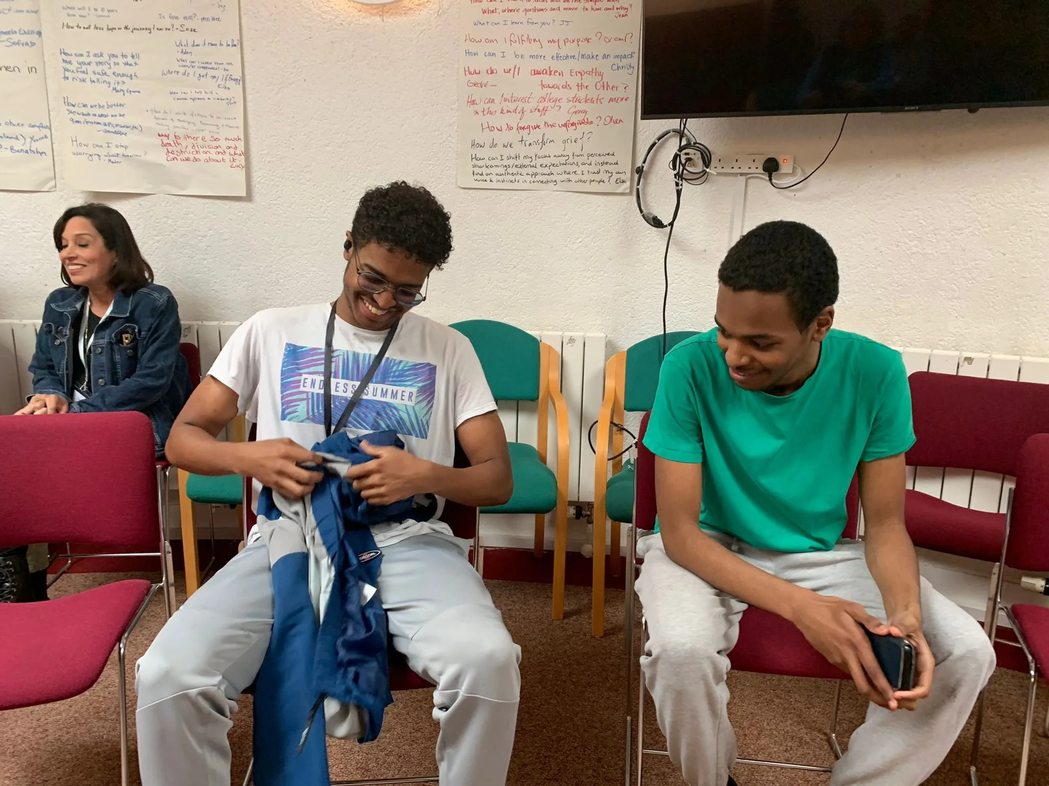 Three people seated in a circle, two men in the foreground smiling and looking down at an object or clothing, woman in background smiling, papers with writing on the wall, and a TV mounted on the wall behind them.