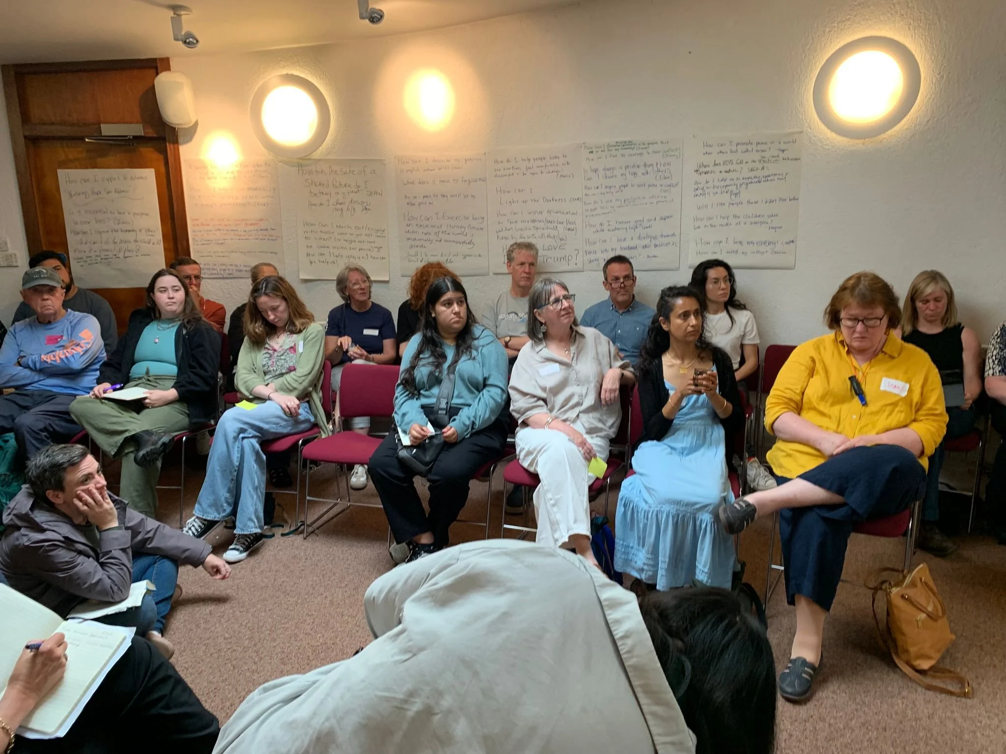 A group of people seated in a room, attentively listening to a presentation or discussion. The room has white walls with large sheets of paper containing handwritten notes taped to the wall.
