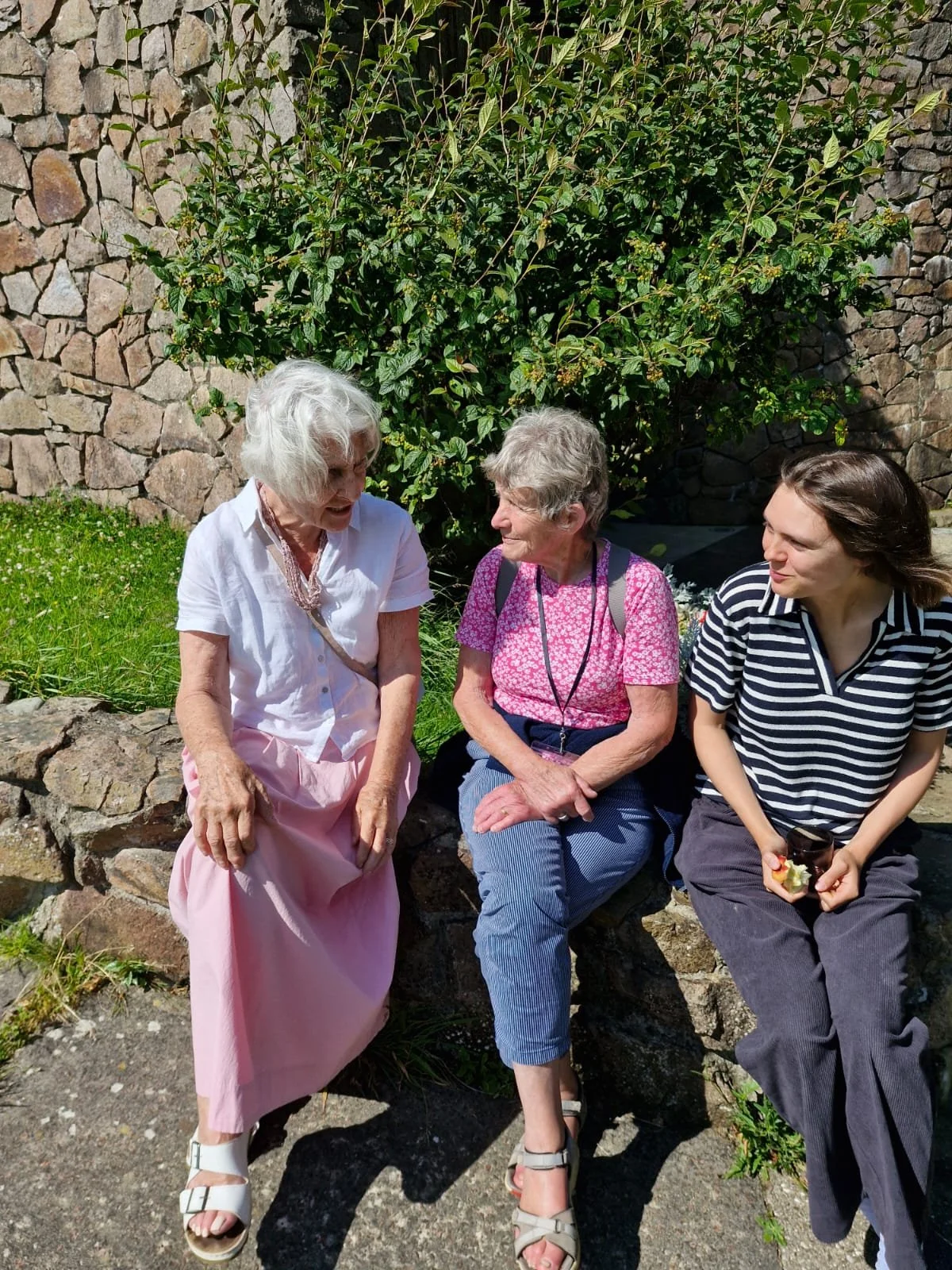 Three women sitting on a stone bench outside, engaged in conversation. 