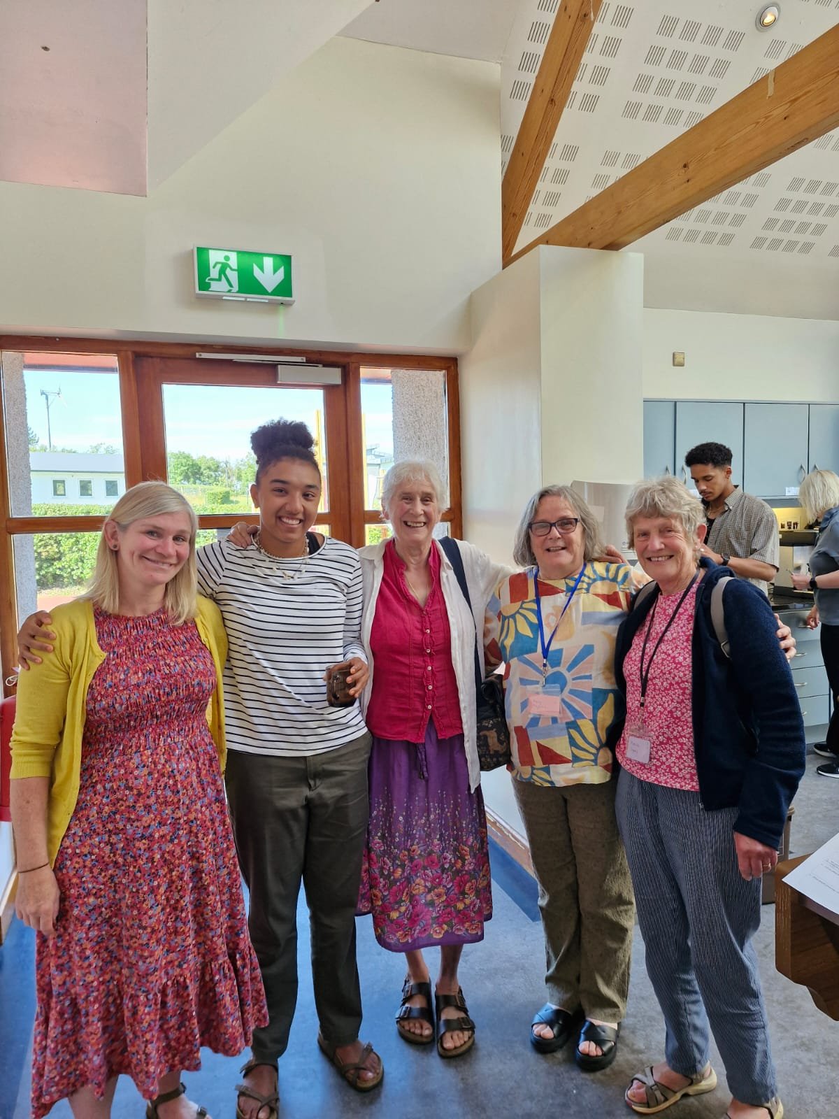 Five women of different ages smiling and standing together indoors. There is a man and a woman working at a counter in the background. Large windows with a view outside, a door with an exit sign above it.