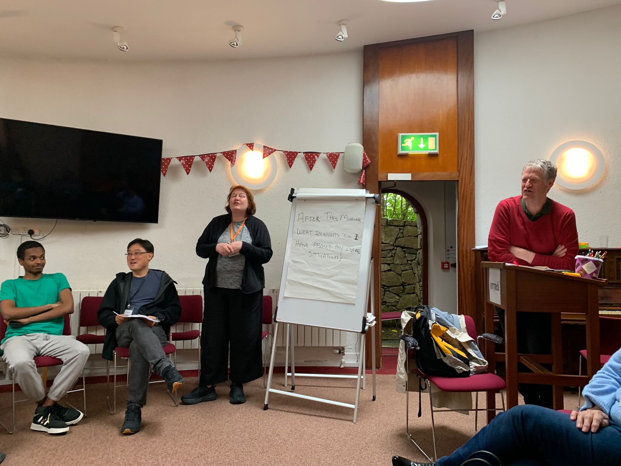 A group of people participating in a workshop or presentation in a room with a large television, a whiteboard, and a wooden high counter. Four individuals are visible, including a woman speaking, two men sitting and listening, and one man standing be