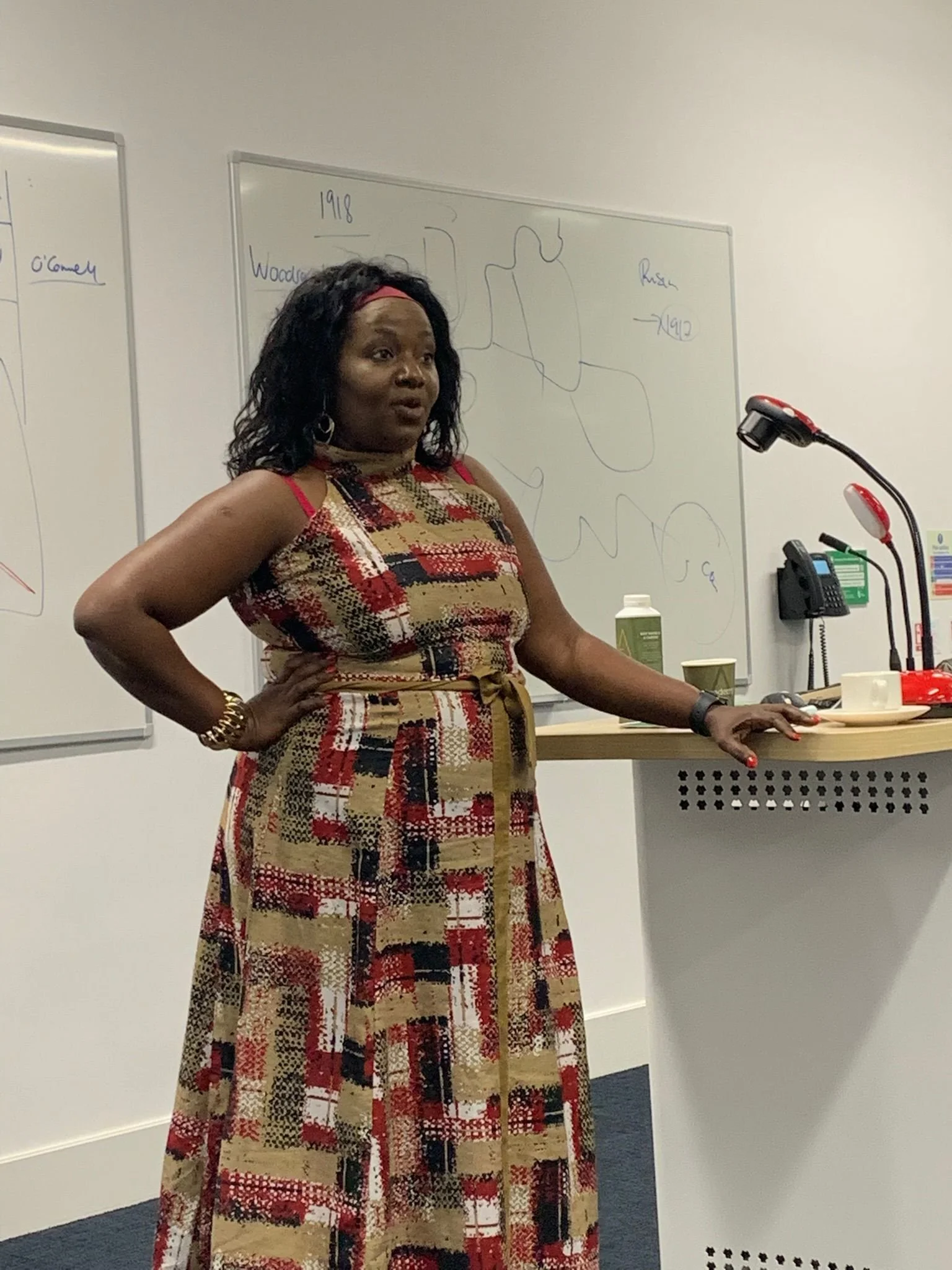 A woman in a patterned dress standing at a lectern in a classroom or conference room, speaking or presenting. There are whiteboards with diagrams and writing in the background.