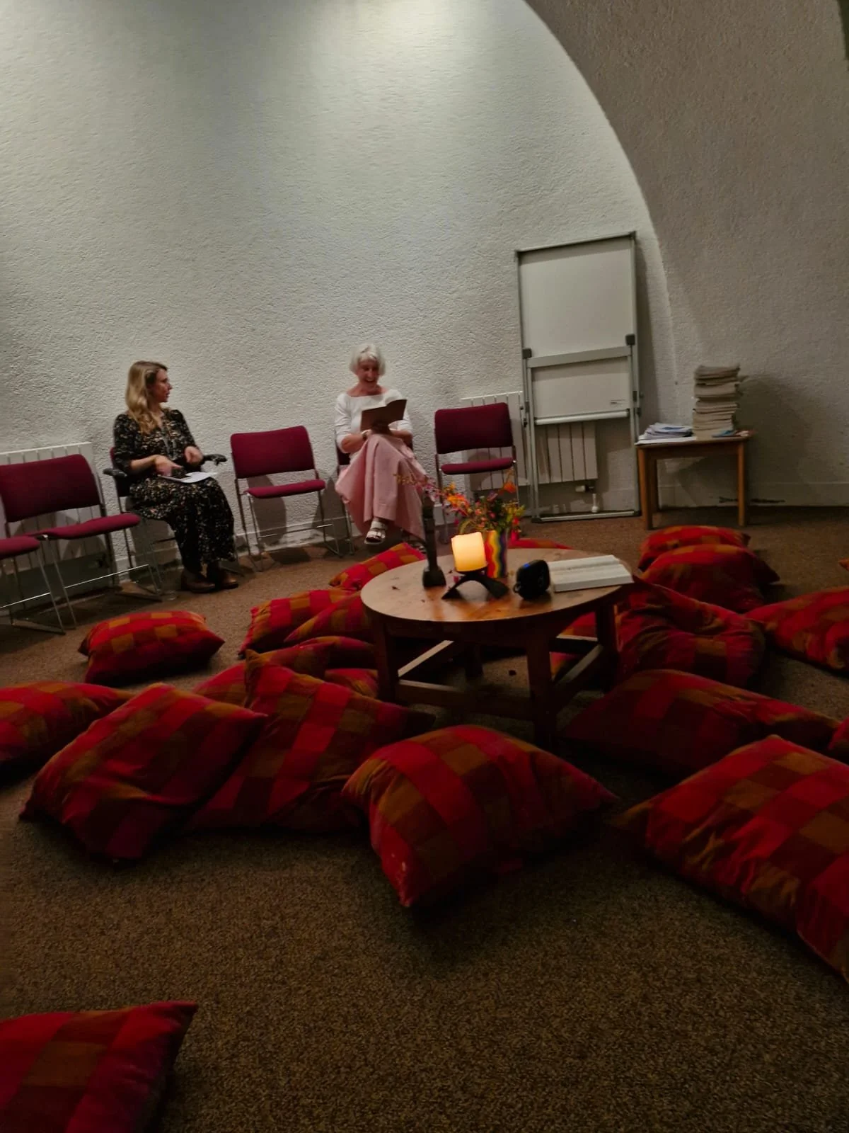 Two women seated on chairs against a white wall, with one reading a book and the other holding papers, in a room with a low table surrounded by red-and-orange checkered cushions, a candle, and books.
