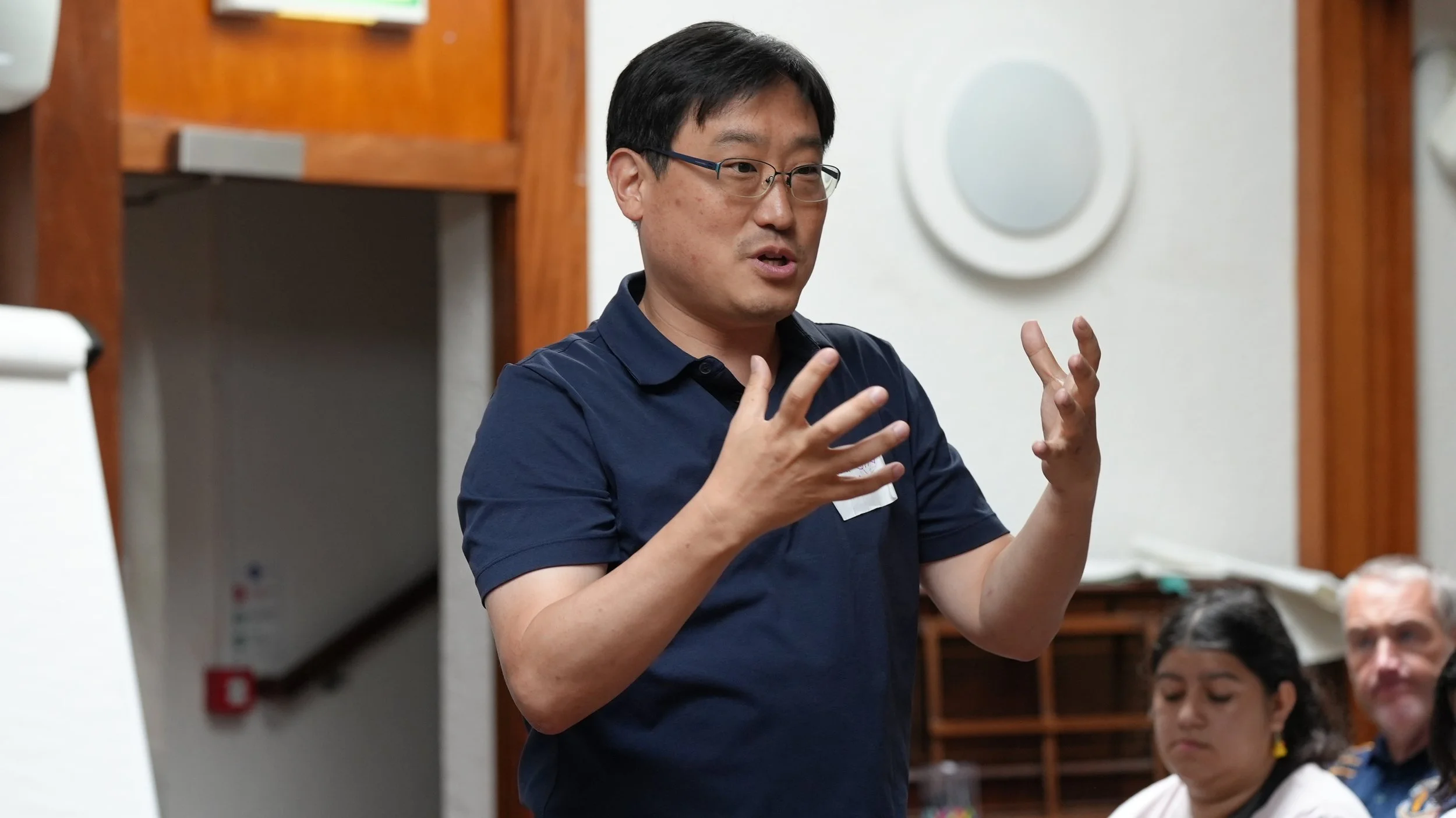A man with glasses and short black hair wearing a navy blue polo shirt speaking or explaining with hand gestures in a room, with a whiteboard and other people listening.