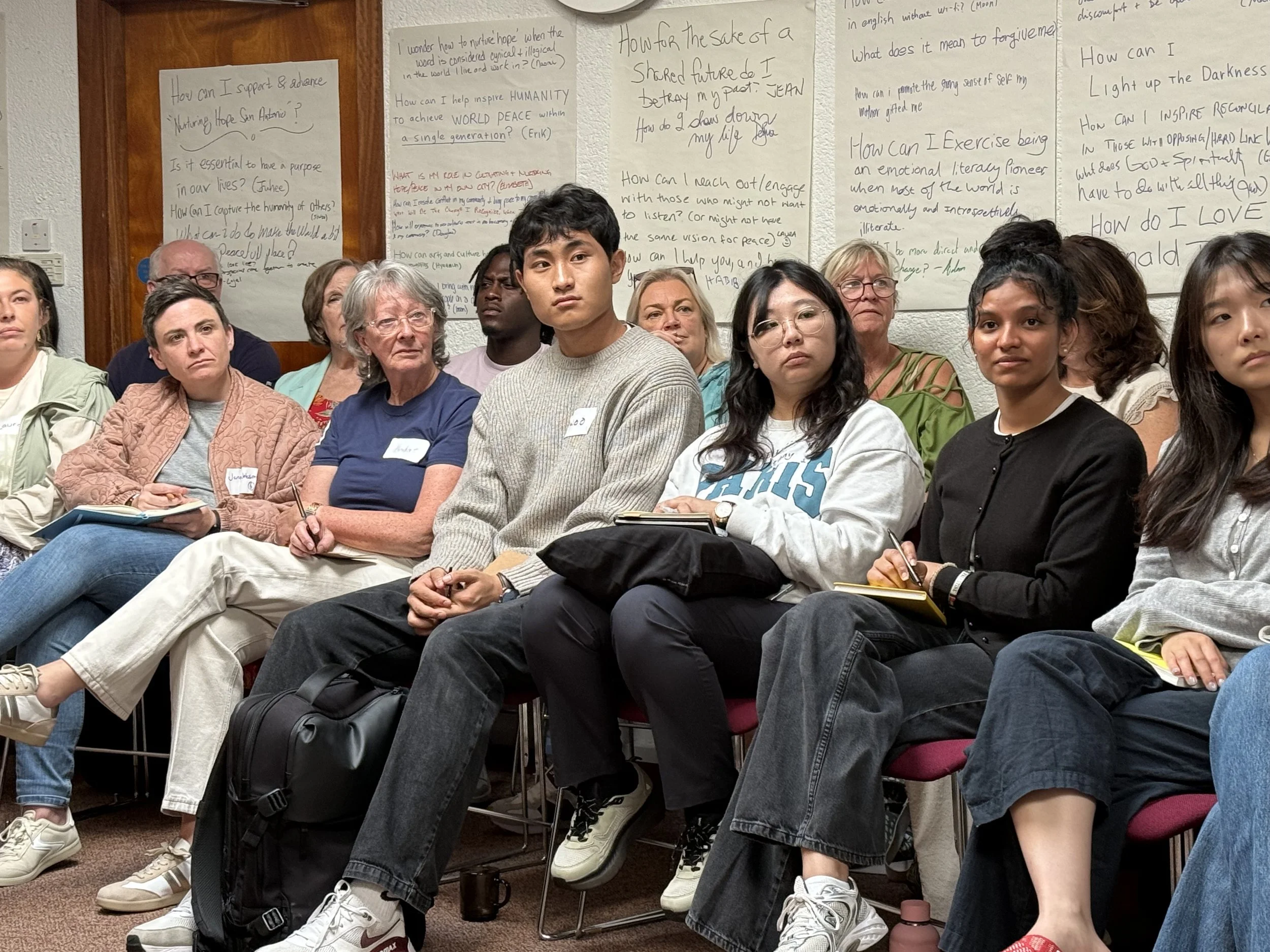 A group of people attending a Nurturing Hope Learning Journey seminar, seated in chairs, taking notes and listening attentively. Behind them are large sheets of paper with handwritten notes and questions about personal growth and social issues.
