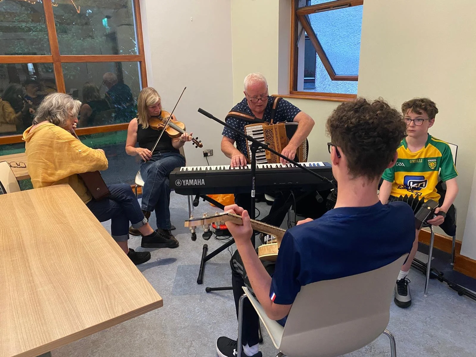 Group of five people playing musical instruments in a room, including a keyboard, accordion, violin, and guitar, with two audience members or other musicians seen through a window.